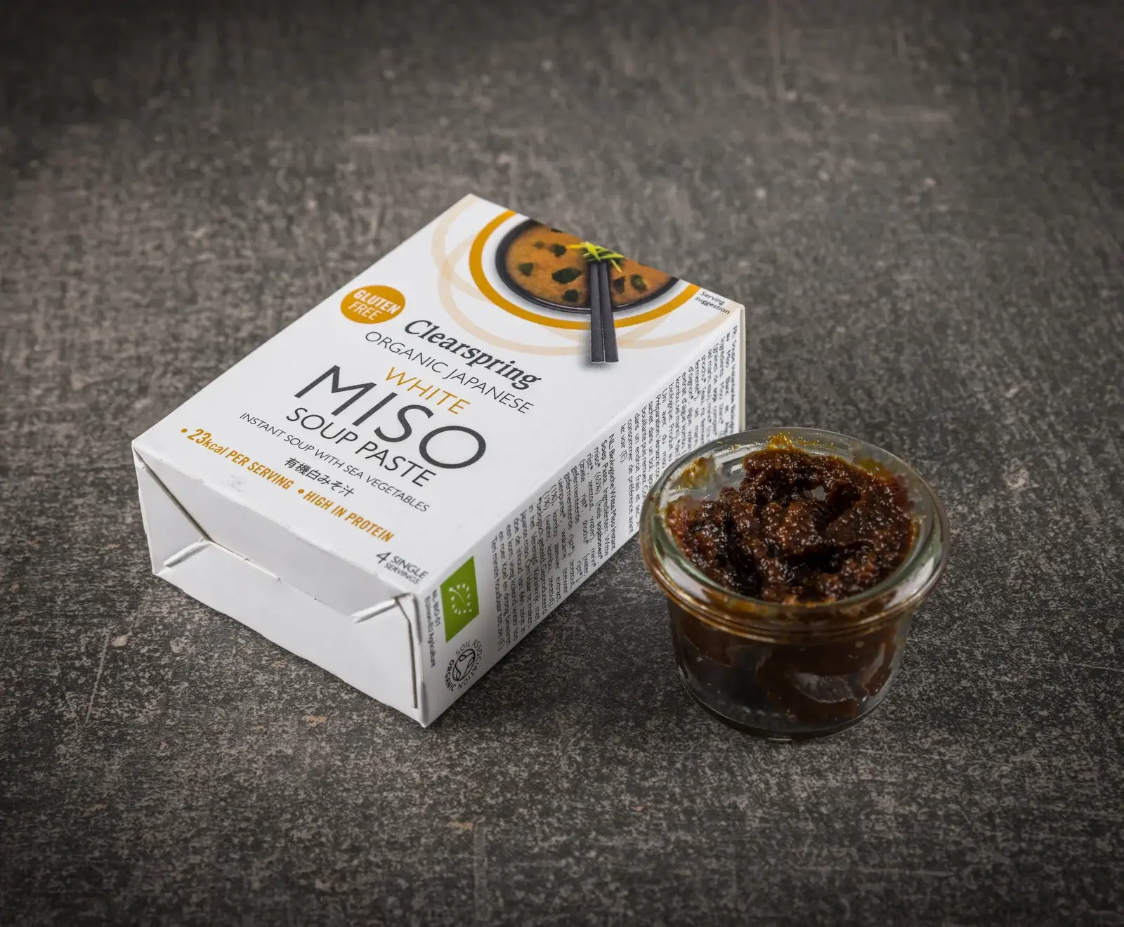 Ein weißer Karton Clearspring Bio Japanische Weiße Miso Suppenpaste, glutenfrei, steht neben einem kleinen Glas mit dunkler, dicker Miso-Paste auf einem dunklen, strukturierten Untergrund.