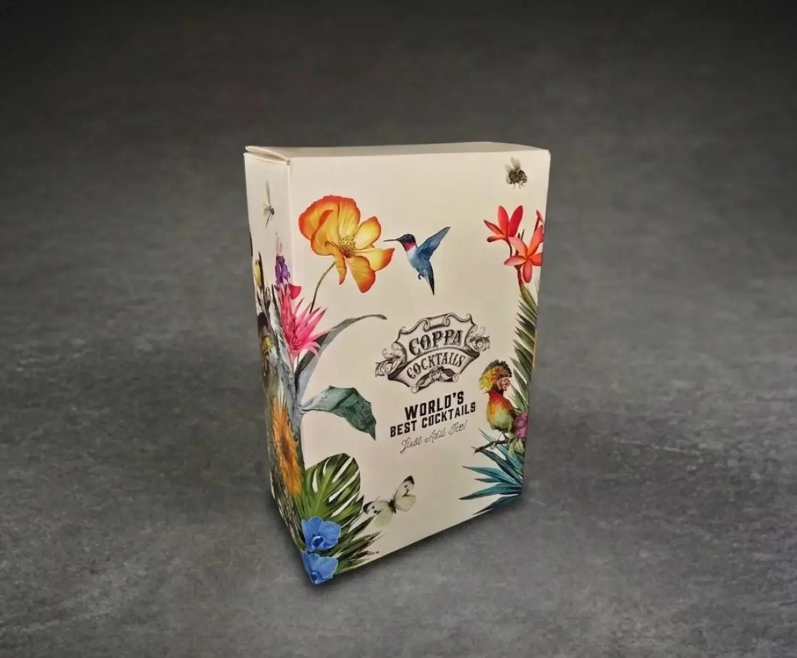 Weiße Pappschachtel für Coppa Cocktails mit farbenfrohen tropischen Illustrationen von Blumen, Kolibri, Papagei, Schmetterling und Insekten. Aufschrift: "WORLD'S BEST COCKTAILS". Steht auf dunklem Untergrund.