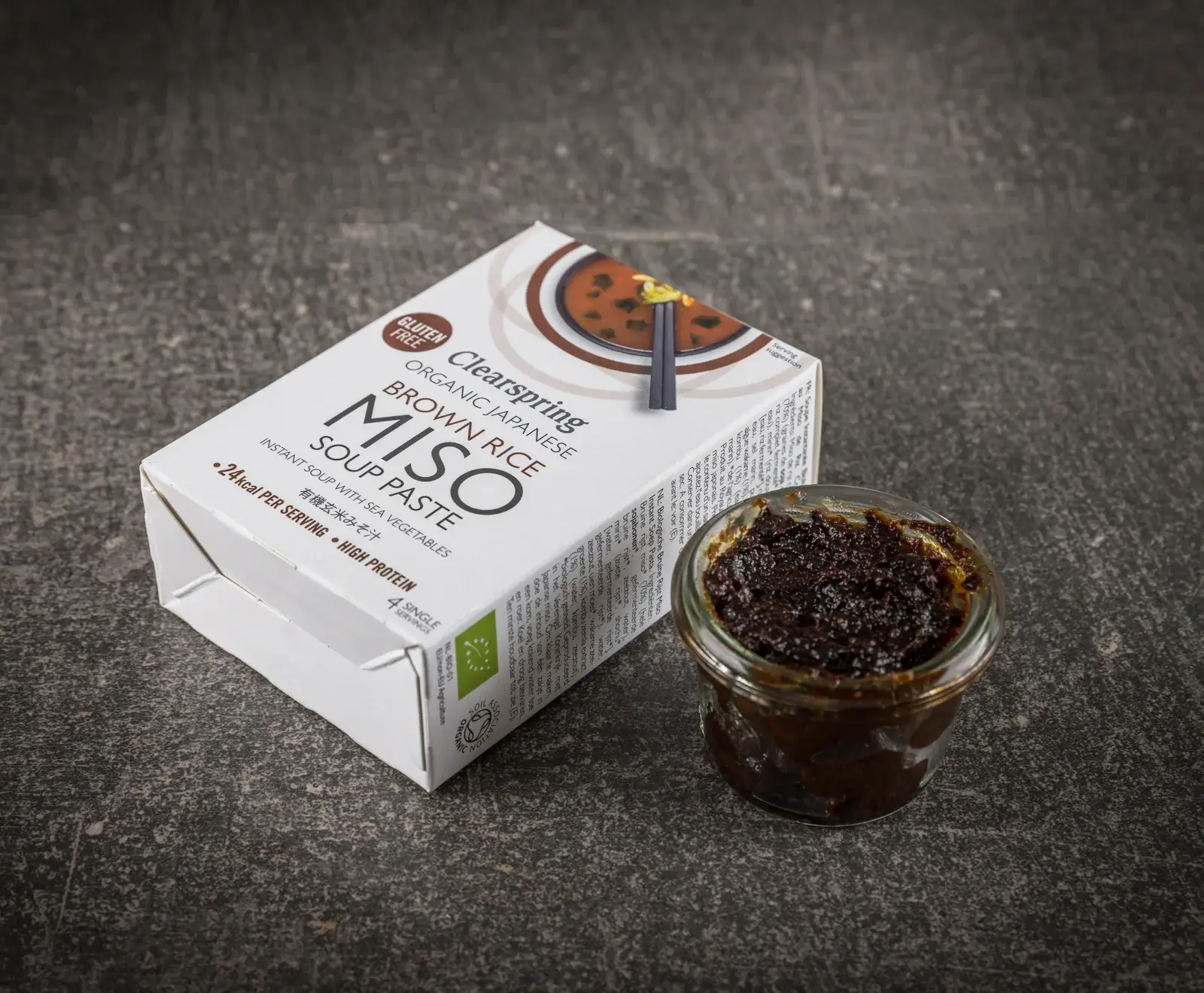 Eine weiße Packung Clearspring Bio Japanische Braunreis Miso Suppenpaste steht neben einem offenen Glas mit dunkler, dickflüssiger Miso-Paste auf einem dunklen Untergrund. Die Packung ist glutenfrei und enthält 4 Portionen.