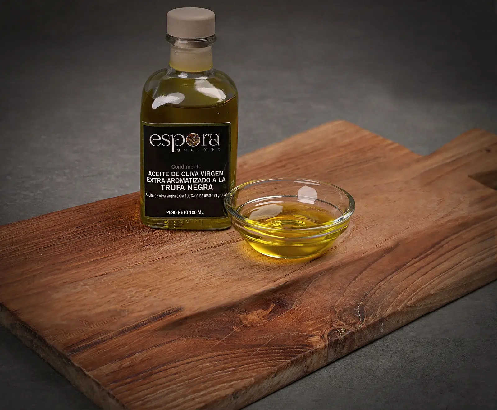 Eine 100-ml-Flasche Espora Gourmet natives Olivenöl extra mit schwarzem Trüffelgeschmack und eine kleine Glasschale mit dem goldenen Öl stehen auf einem rustikalen Holzschneidebrett.