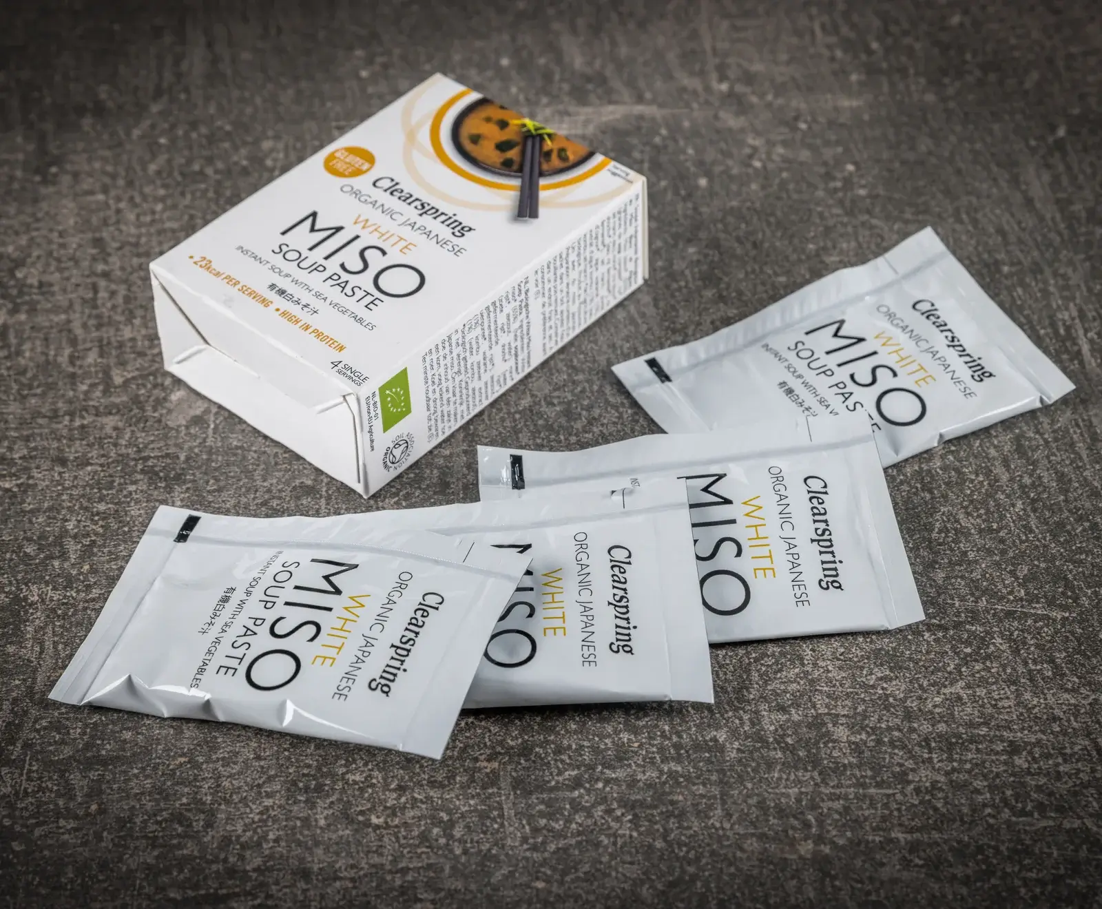 Eine weiße Schachtel Clearspring Bio Japanische Weiße Miso Suppenpaste und vier einzelne weiße Portionsbeutel davon liegen auf einer dunklen, strukturierten Oberfläche.