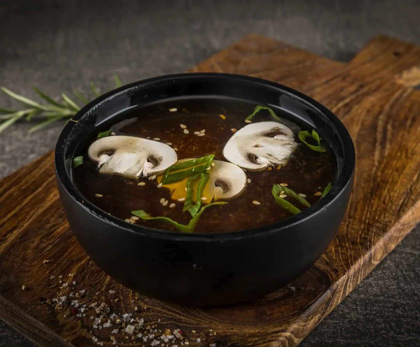 Eine schwarze Schale mit dunkler Pilzsuppe, garniert mit weißen Champignonscheiben, grünen Zwiebeln und Sesamsamen, steht auf einem rustikalen Holzbrett mit Salz und Pfeffer.