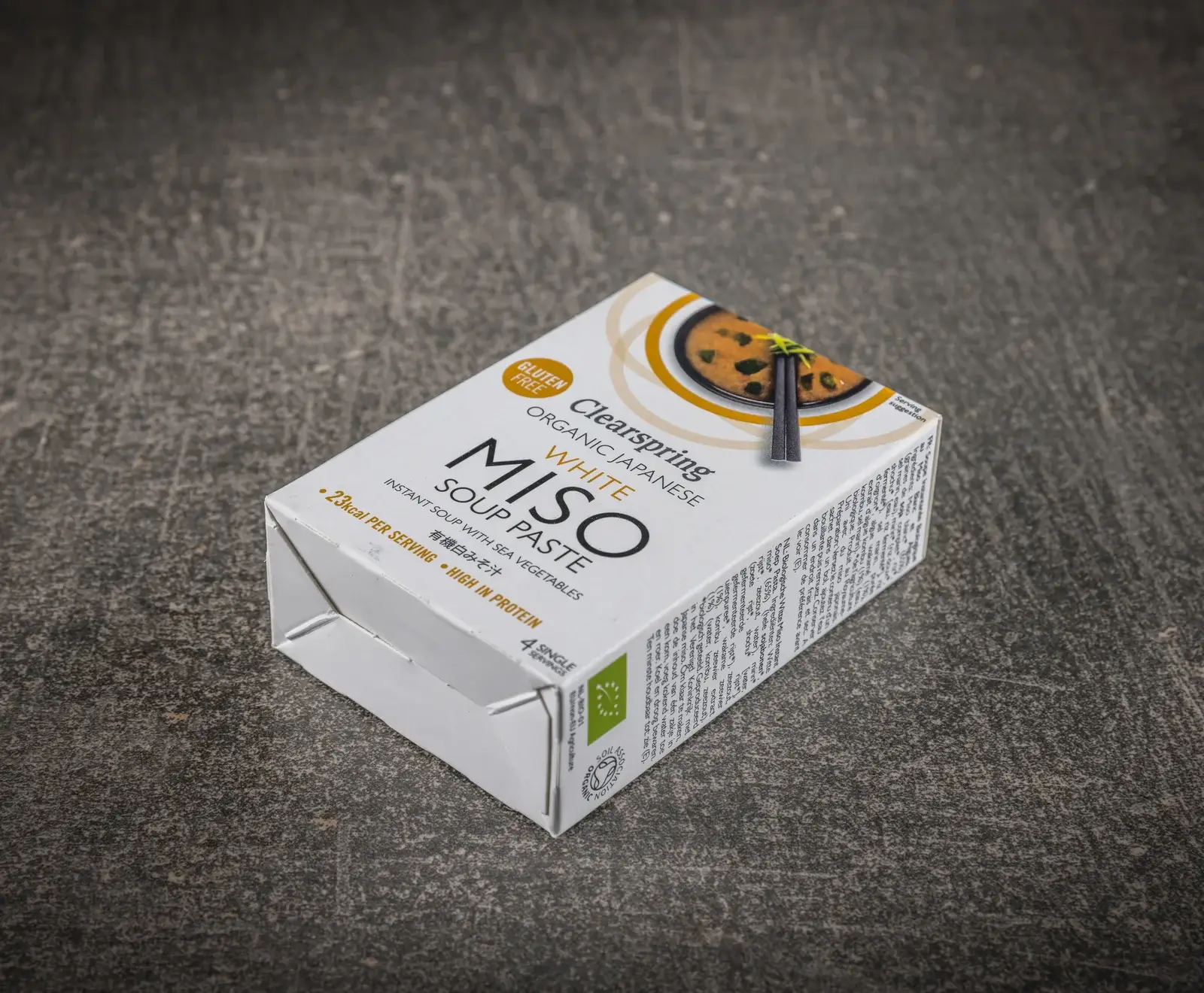 Eine weiße Packung Clearspring Bio Japanische Weiße Miso Suppenpaste, glutenfrei und proteinreich, steht schräg auf einer dunklen Oberfläche. Die Vorderseite zeigt eine Schale Miso-Suppe mit Essstäbchen.