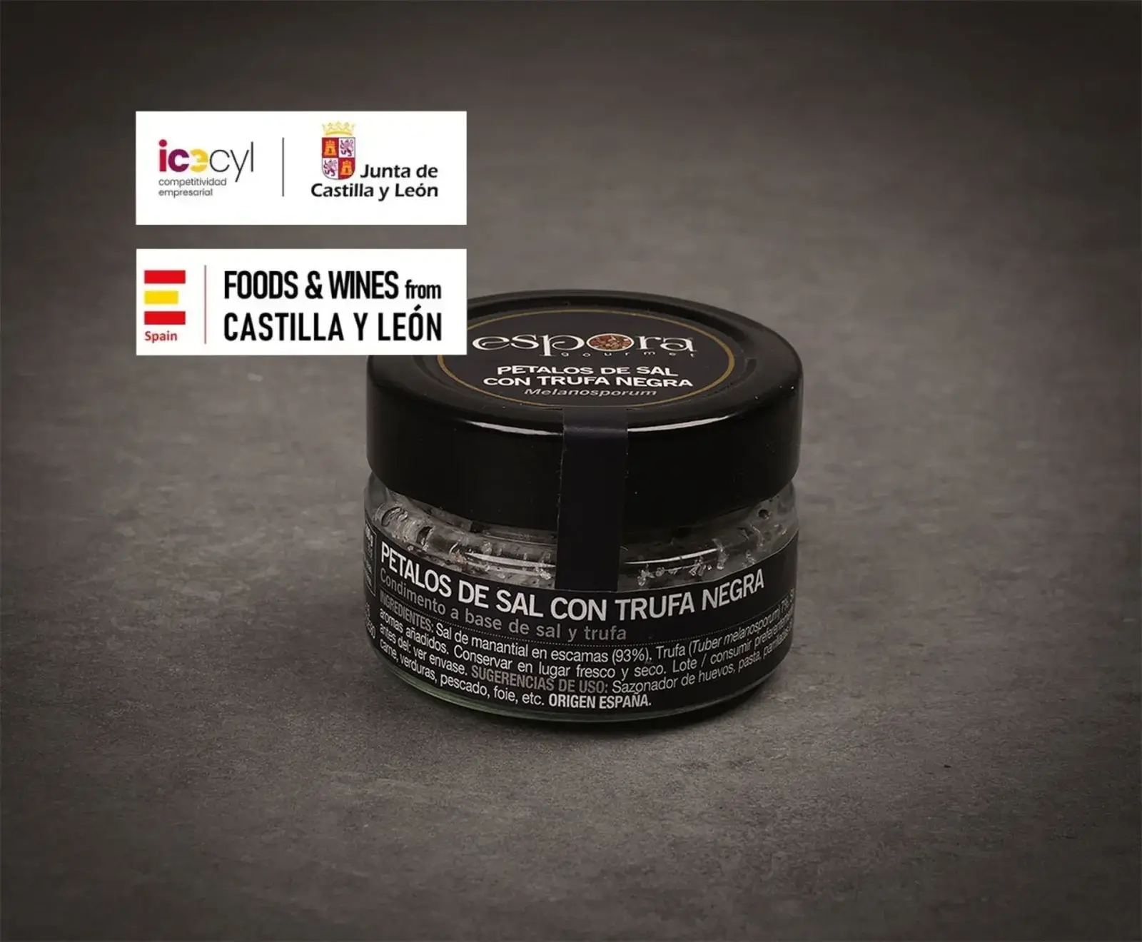 Ein kleines Glasgefäß mit schwarzem Deckel, gefüllt mit schwarzen Trüffelsalzflocken der Marke Espora Gourmet. Oben links sind Logos von "Junta de Castilla y León" und "Foods & Wines from Castilla y León" zu sehen.