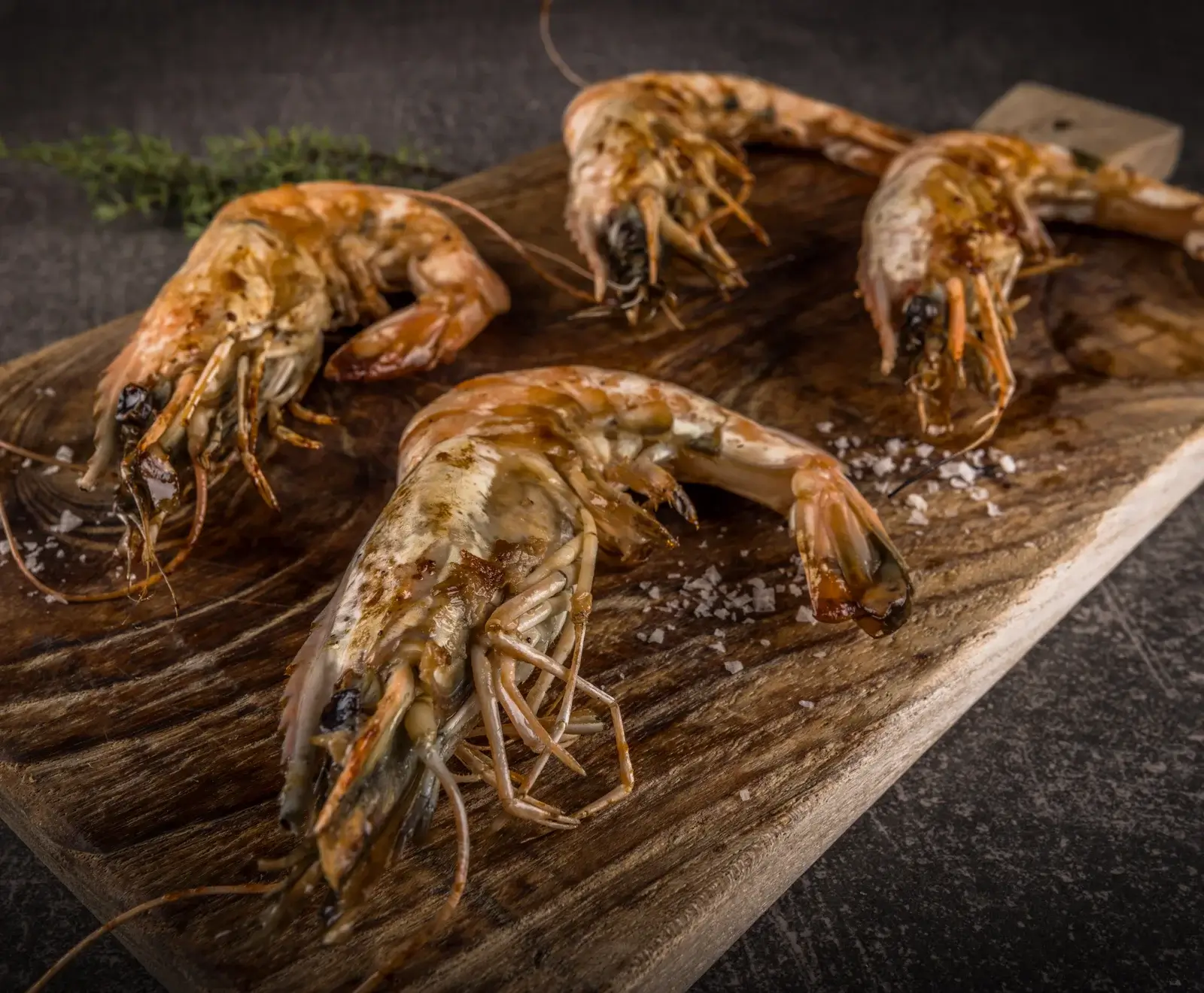 Mehrere goldbraun gegrillte Riesengarnelen mit Schale liegen auf einem rustikalen Holzbrett. Grobes Meersalz ist verstreut, ein Kräuterzweig im dunklen Hintergrund. Stimmungsvolle Beleuchtung.
