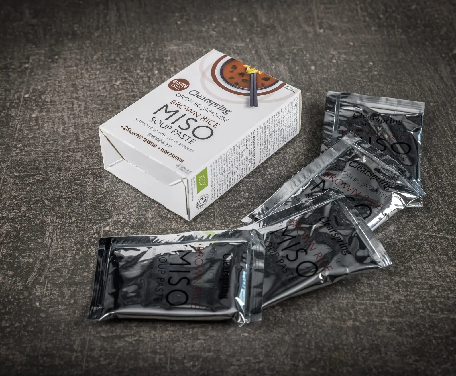 Eine weiße Schachtel Clearspring Bio Japanische Brauner Reis Miso Suppenpaste mit der Aufschrift "Glutenfrei" und "4 Einzelportionen" liegt auf einer dunklen Oberfläche. Daneben liegen vier silberne Einzelportionsbeutel der Suppenpaste.