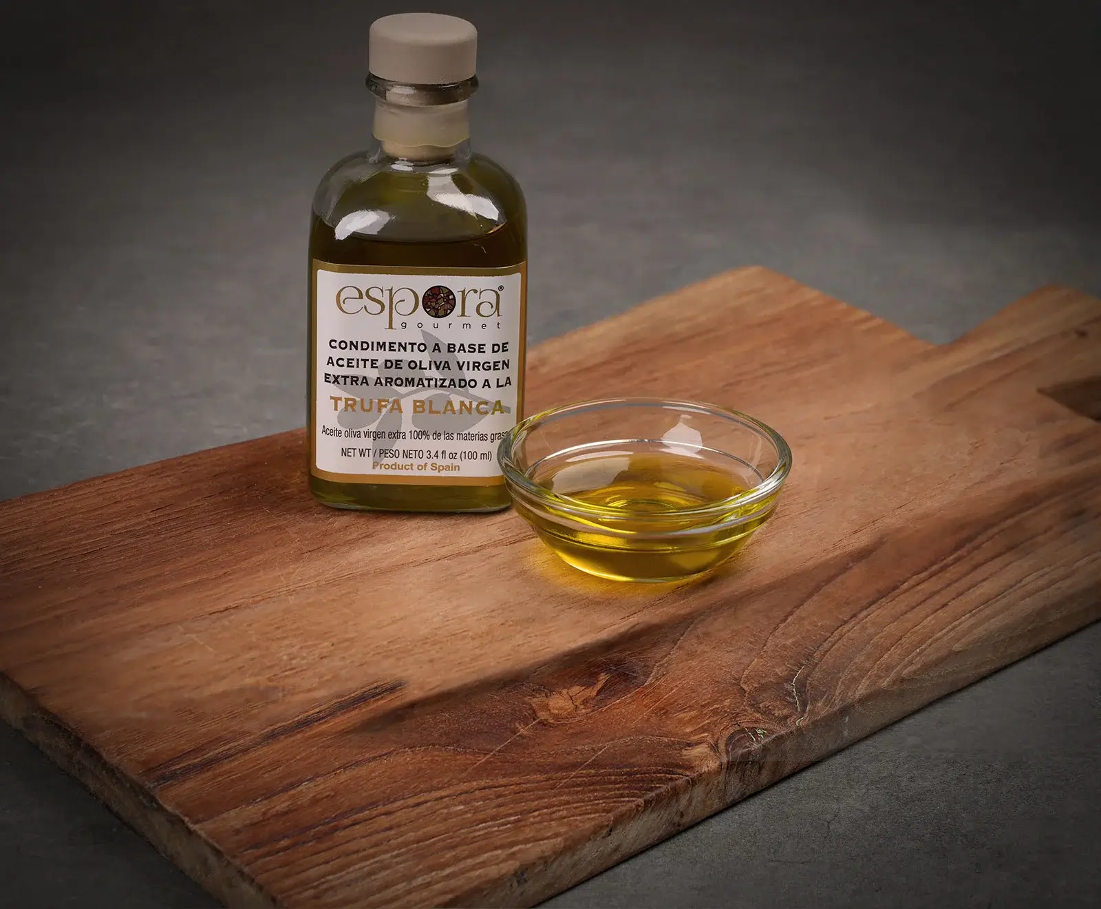 Eine Flasche Espora Gourmet natives Olivenöl extra mit weißem Trüffelgeschmack und eine kleine Glasschale mit dem goldenen Öl stehen auf einem rustikalen Holzschneidebrett vor dunklem Hintergrund.