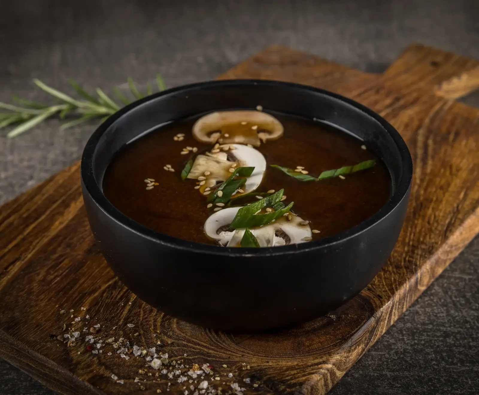 Eine schwarze Schale mit dunkler Pilzsuppe, garniert mit Champignonscheiben, grünen Kräutern und Sesamkörnern, steht auf einem rustikalen Holzbrett. Im Hintergrund Rosmarin, im Vordergrund Salz und Pfeffer.