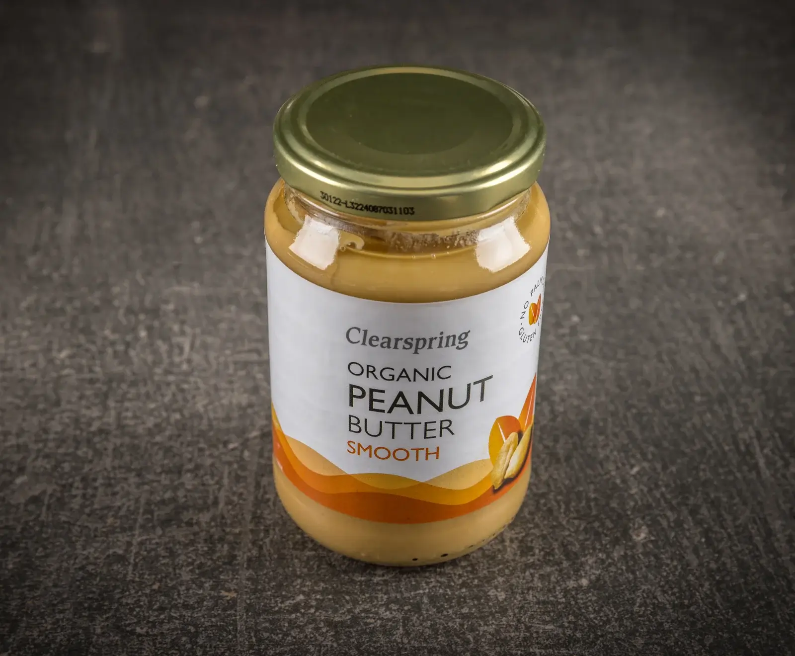 Ein Glas Clearspring Bio Erdnussbutter Smooth mit goldenem Deckel, auf dunklem Hintergrund. Das Etikett zeigt den Markennamen, die Sorte und Erdnüsse, sowie Hinweise auf palmöl- und glutenfrei.