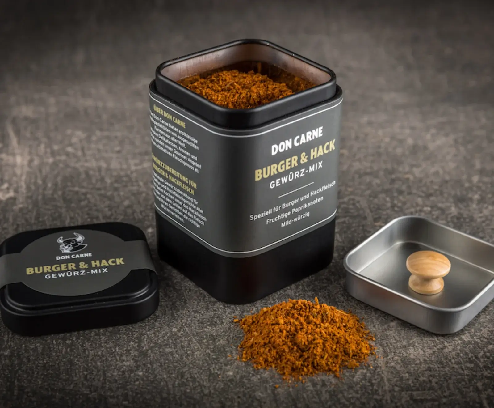 Ein offenes schwarzes Gewürzdosen-Set von Don Carne "Burger & Hack Gewürz-Mix" auf dunklem Untergrund. Die Dose enthält das rotbraune Gewürz, daneben liegt ein Haufen davon, der Deckel und eine kleine Metallschale mit Holzgriff.