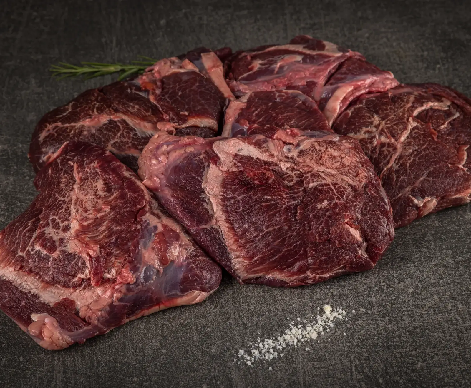 Mehrere Stücke rohes, marmoriertes Rindfleisch liegen auf einem dunklen Untergrund, begleitet von einem Rosmarinzweig und grobem Salz.