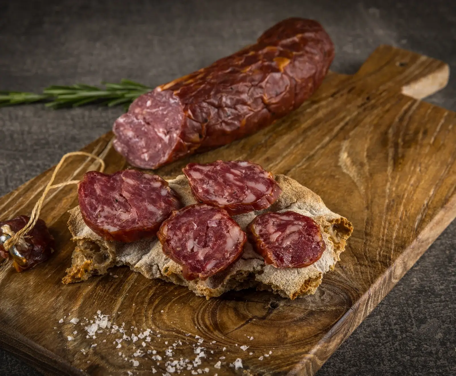 Eine angeschnittene Salami und mehrere Scheiben davon auf einem Stück Fladenbrot, garniert mit grobem Salz und einem Rosmarinzweig, auf einem rustikalen Holzschneidebrett.