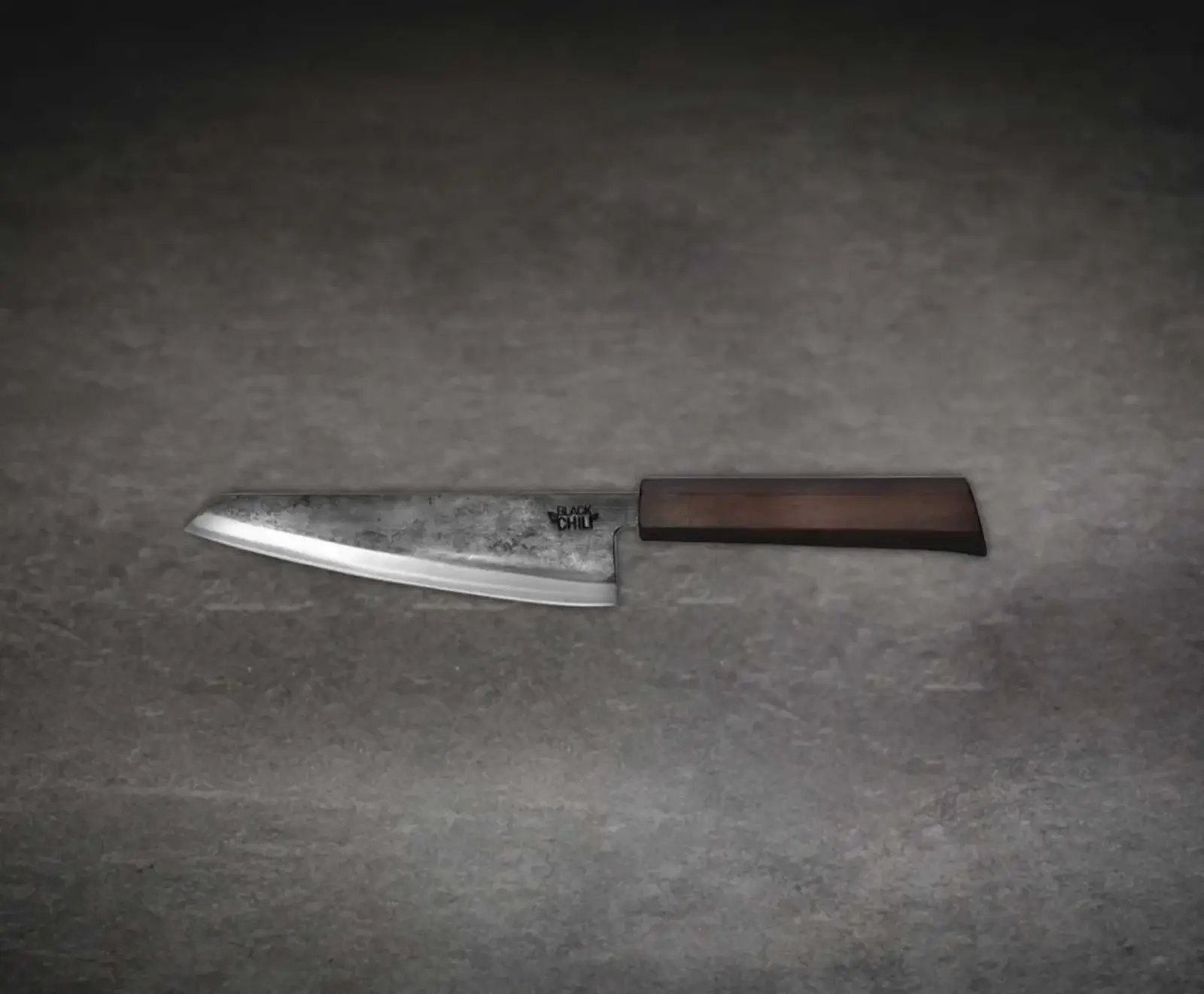 Ein rustikales Kochmesser mit breiter, dunkler, strukturierter Klinge und scharfer Schneide liegt auf einem grauen Untergrund. Der Griff ist aus dunklem Holz. Auf der Klinge ist das Logo "BLACK CHILI" zu sehen.