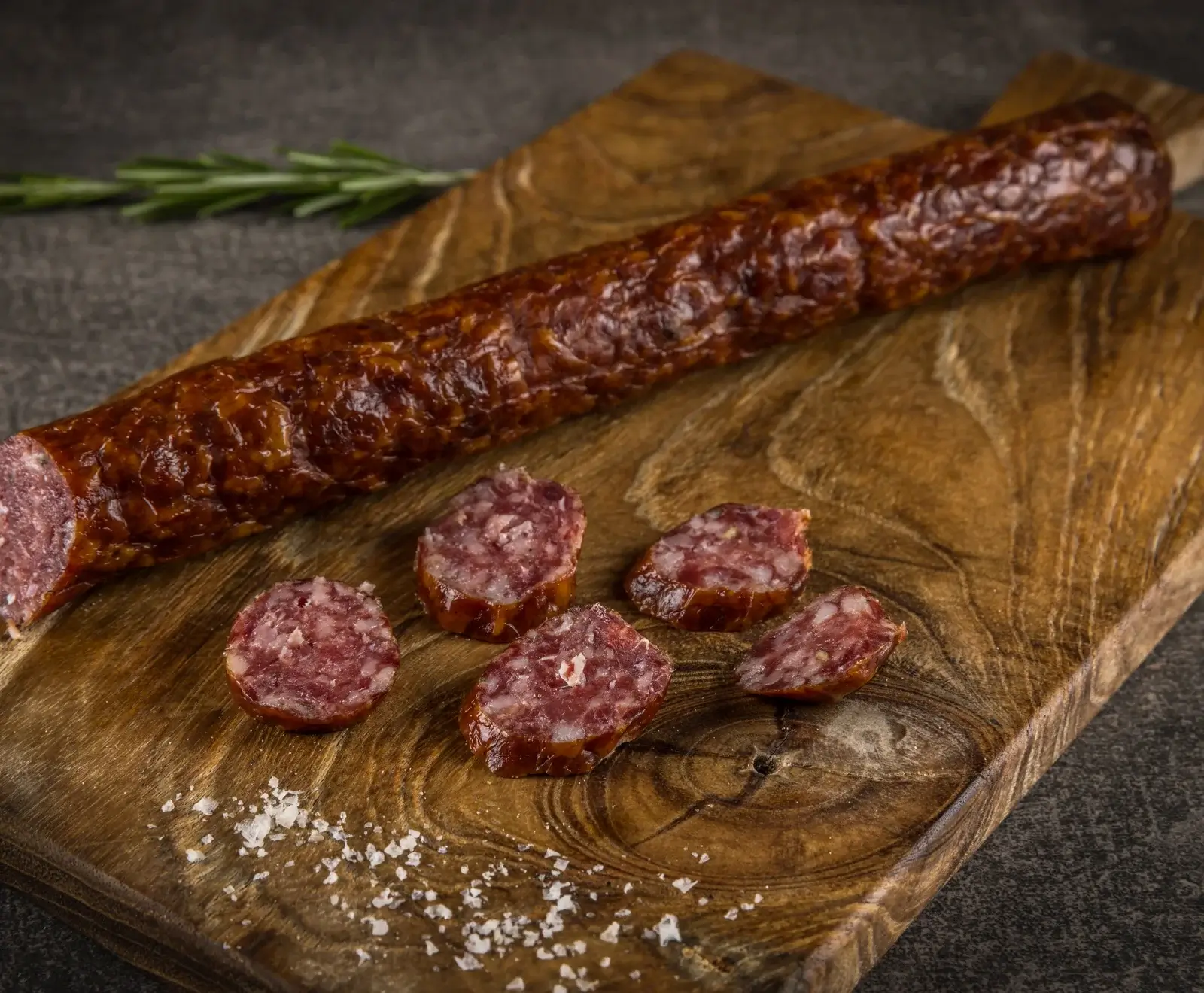Eine lange, dunkelrote Rohwurst liegt auf einem rustikalen Holzbrett, daneben mehrere angeschnittene Scheiben und grobes Salz. Im Hintergrund ist ein Rosmarinzweig zu sehen.