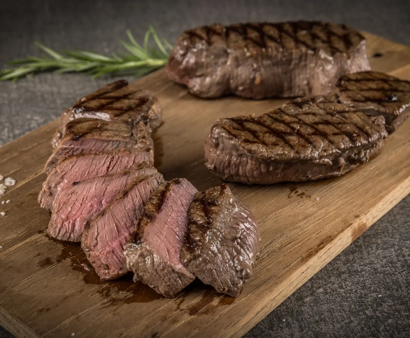 Drei gegrillte Steaks auf einem Holzbrett. Eines ist in Scheiben geschnitten und zeigt eine rosige Mitte. Die Steaks haben Grillstreifen. Im Hintergrund Rosmarin und grobes Salz.