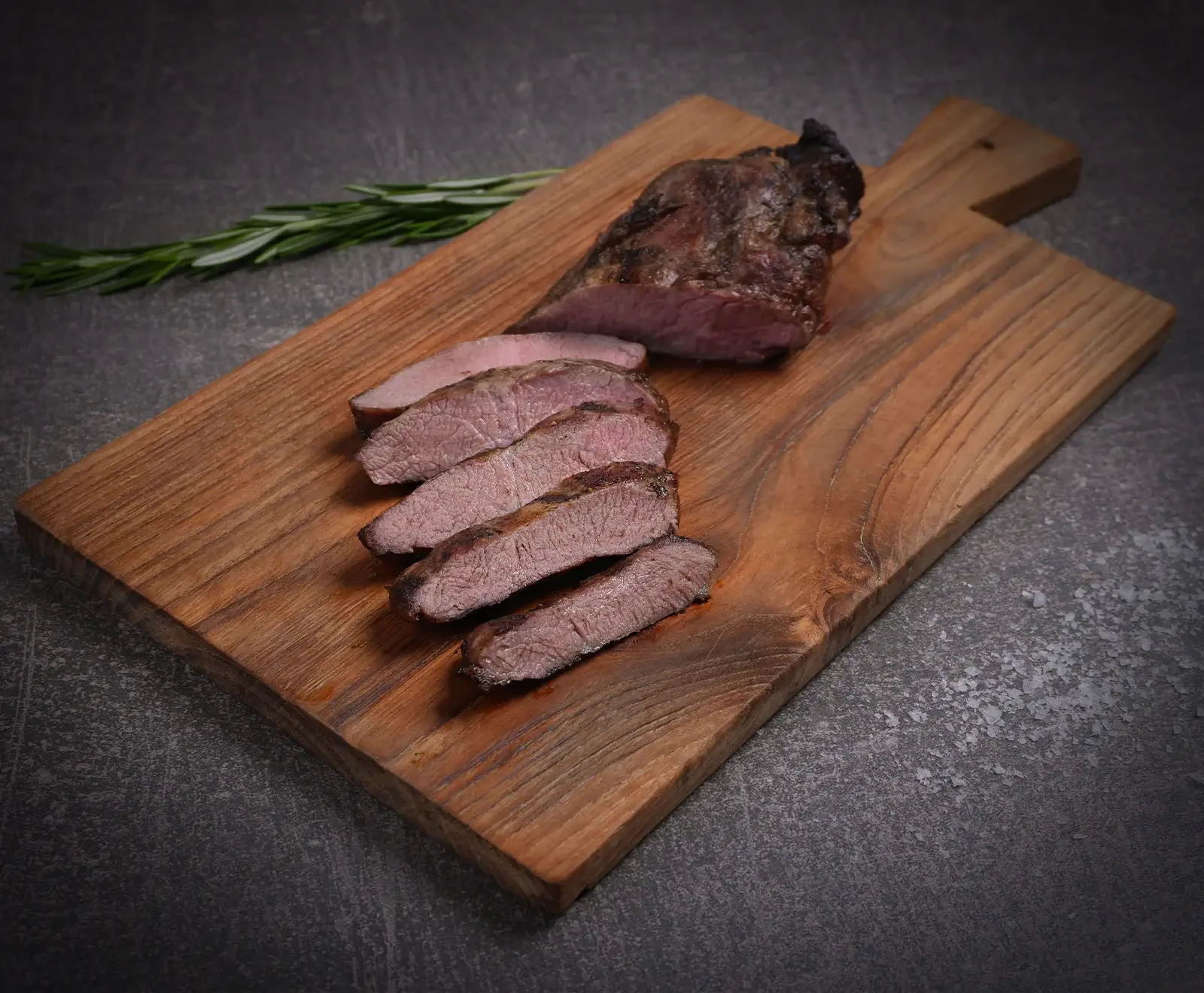 Ein auf einem rustikalen Holzschneidebrett angerichtetes, rosa gebratenes Stück Fleisch, teilweise in fünf Scheiben geschnitten, daneben ein frischer Rosmarinzweig. Der Hintergrund ist dunkelgrau.