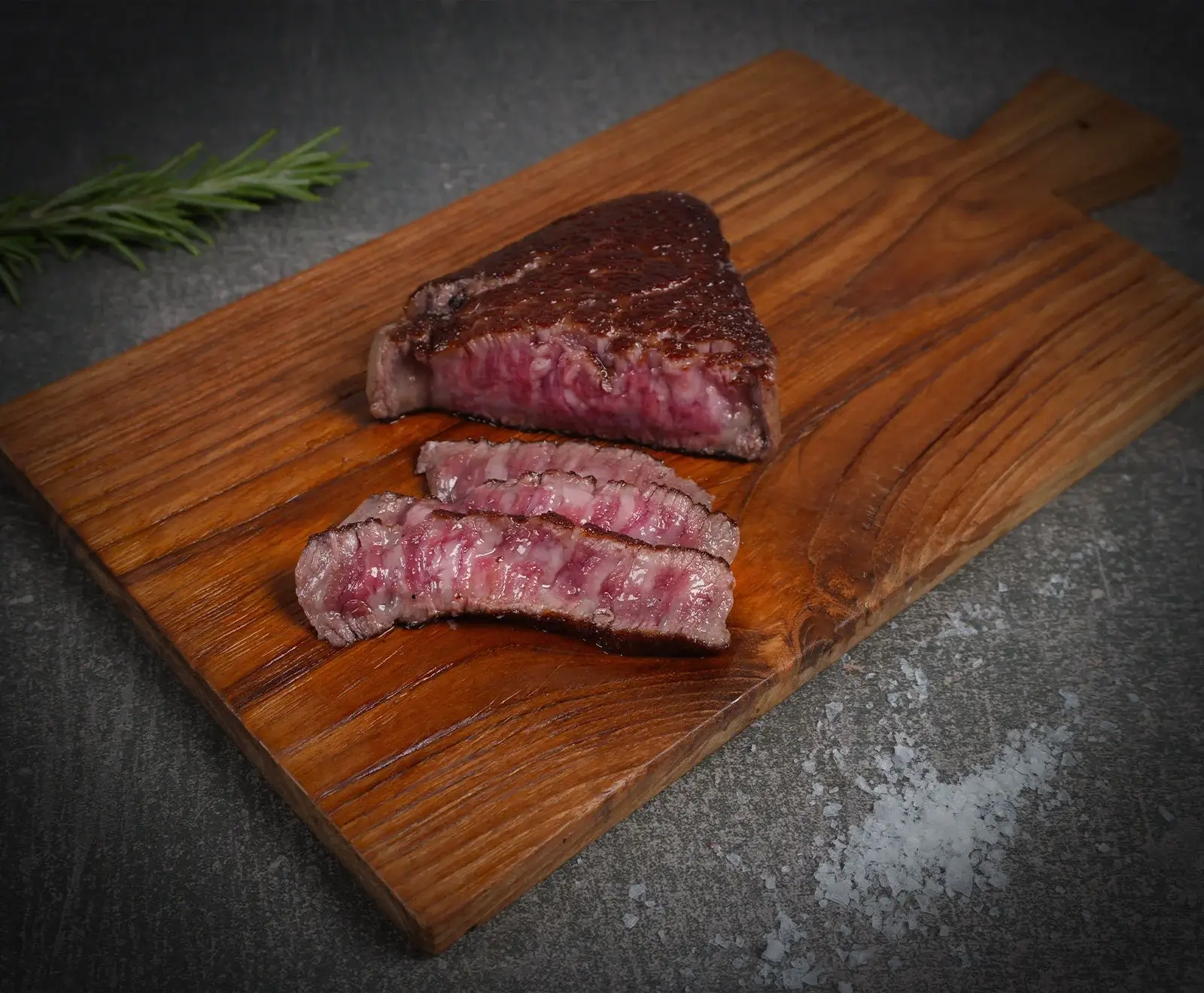 Ein perfekt gebratenes, aufgeschnittenes Steak mit rosa Kern und Marmorierung auf einem Holzbrett. Daneben liegen Rosmarin und grobes Meersalz auf dunklem Untergrund.