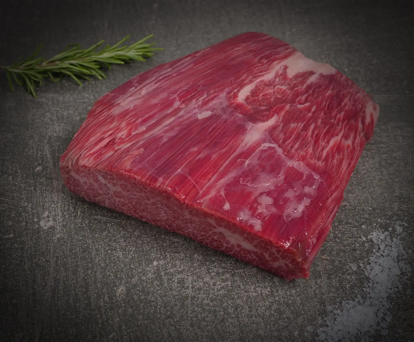 Ein rohes Stück rotes Rindfleisch mit feiner Marmorierung und sichtbaren Fettadern liegt auf einer dunklen Steinplatte. Daneben ein Rosmarinzweig und grobes Salz.