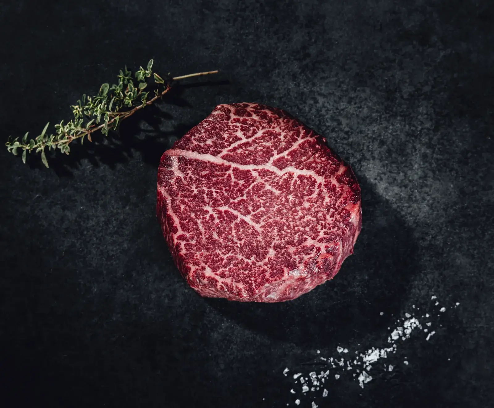 Ein rohes, stark marmoriertes Rinderfiletsteak liegt auf einer dunklen Oberfläche, daneben ein Thymianzweig und grobe Salzkristalle.