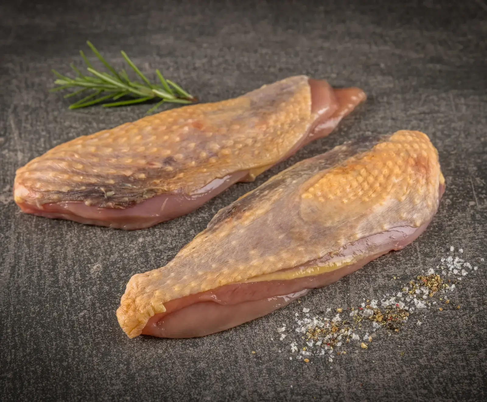 Zwei rohe Entenbrustfilets mit Haut auf dunklem Schiefer, garniert mit einem Rosmarinzweig und einer Prise grobem Salz und Pfeffer.