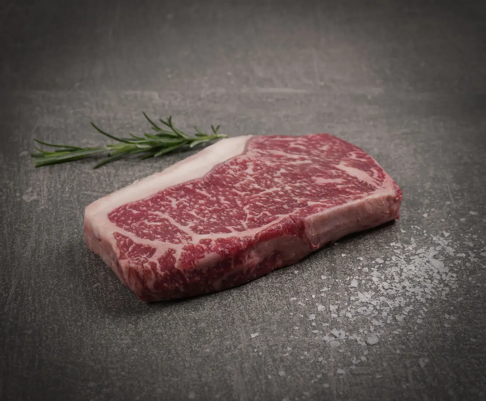 Rohes, stark marmoriertes Rindersteak mit Fettrand auf dunklem Untergrund, daneben ein Rosmarinzweig und grobes Meersalz.