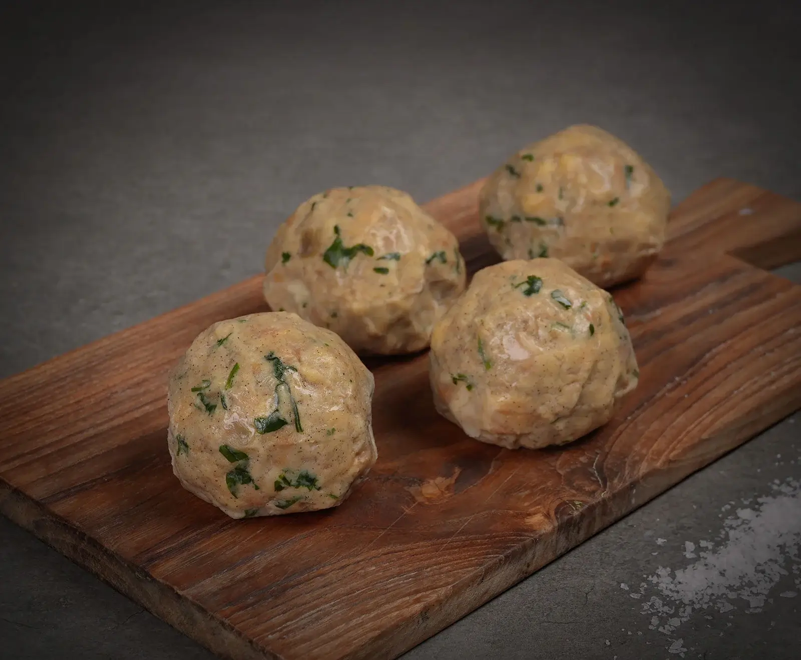 Vier rohe, runde Knödel mit sichtbaren grünen Kräutern auf einem Holzbrett. Der dunkle Hintergrund zeigt rechts unten verstreute Salzkristalle.