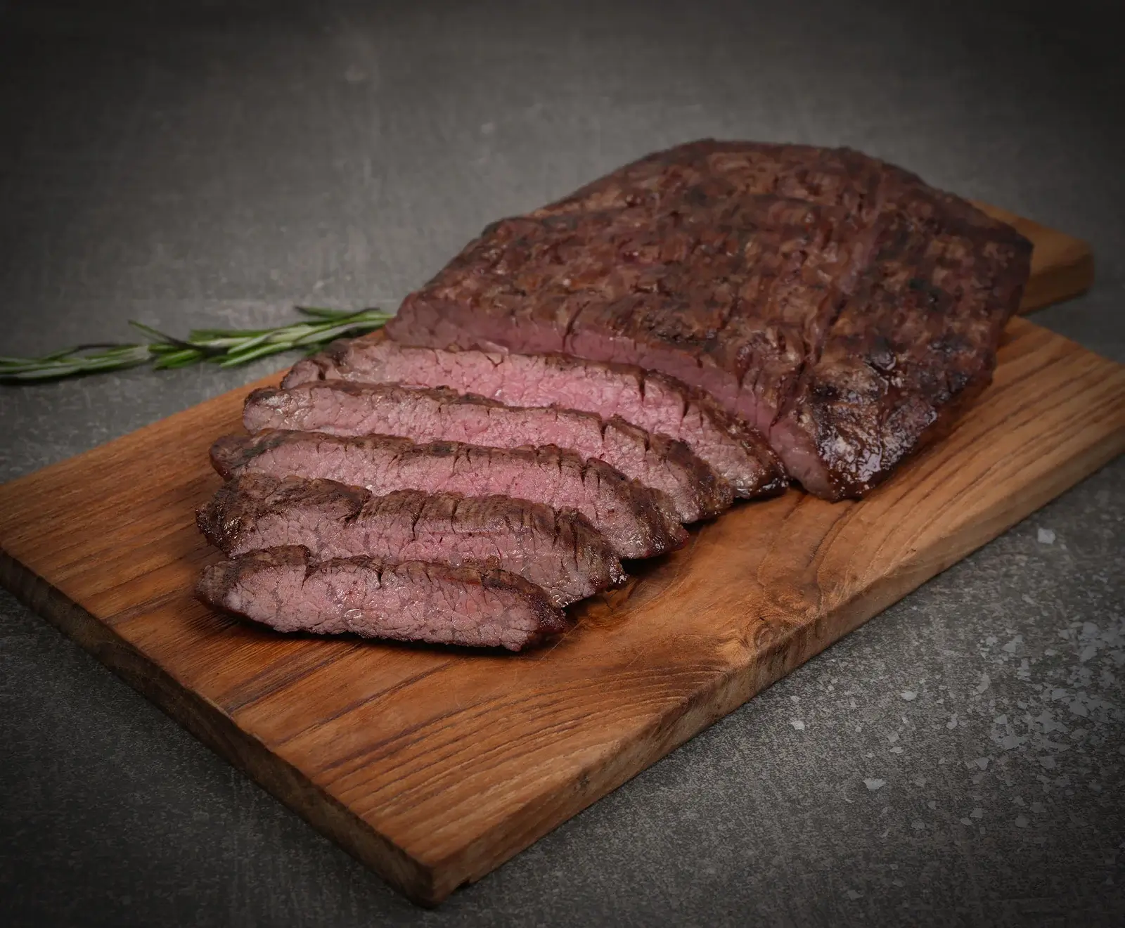 Ein aufgeschnittenes, medium-rare gegrilltes Steak auf einem rustikalen Holzbrett, garniert mit Rosmarin und grobem Salz auf dunklem Hintergrund.