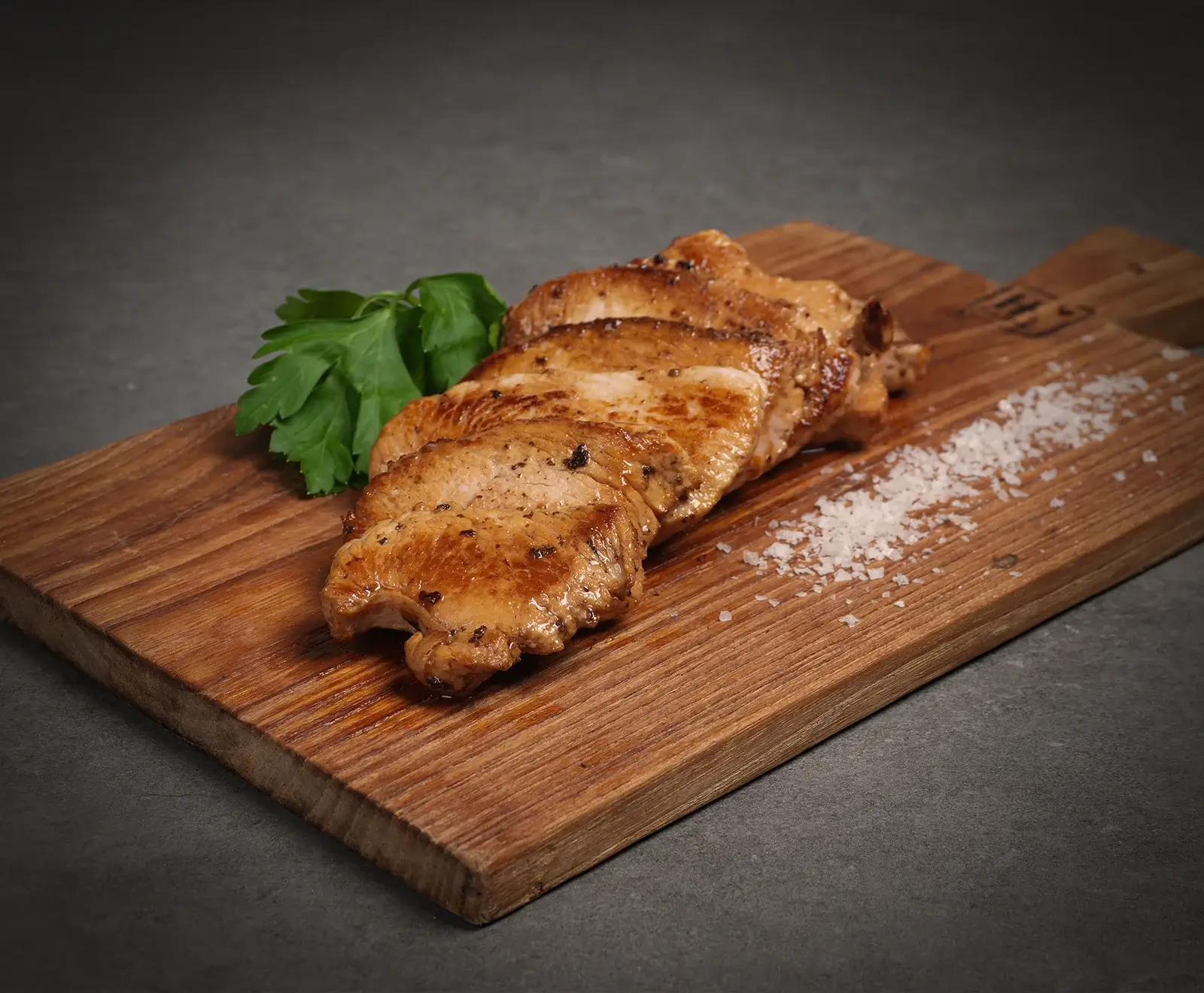 Nahaufnahme von goldbraun gebratenen Fleischscheiben, garniert mit Petersilie und grobem Meersalz, auf einem rustikalen Holzbrett vor dunklem Hintergrund.