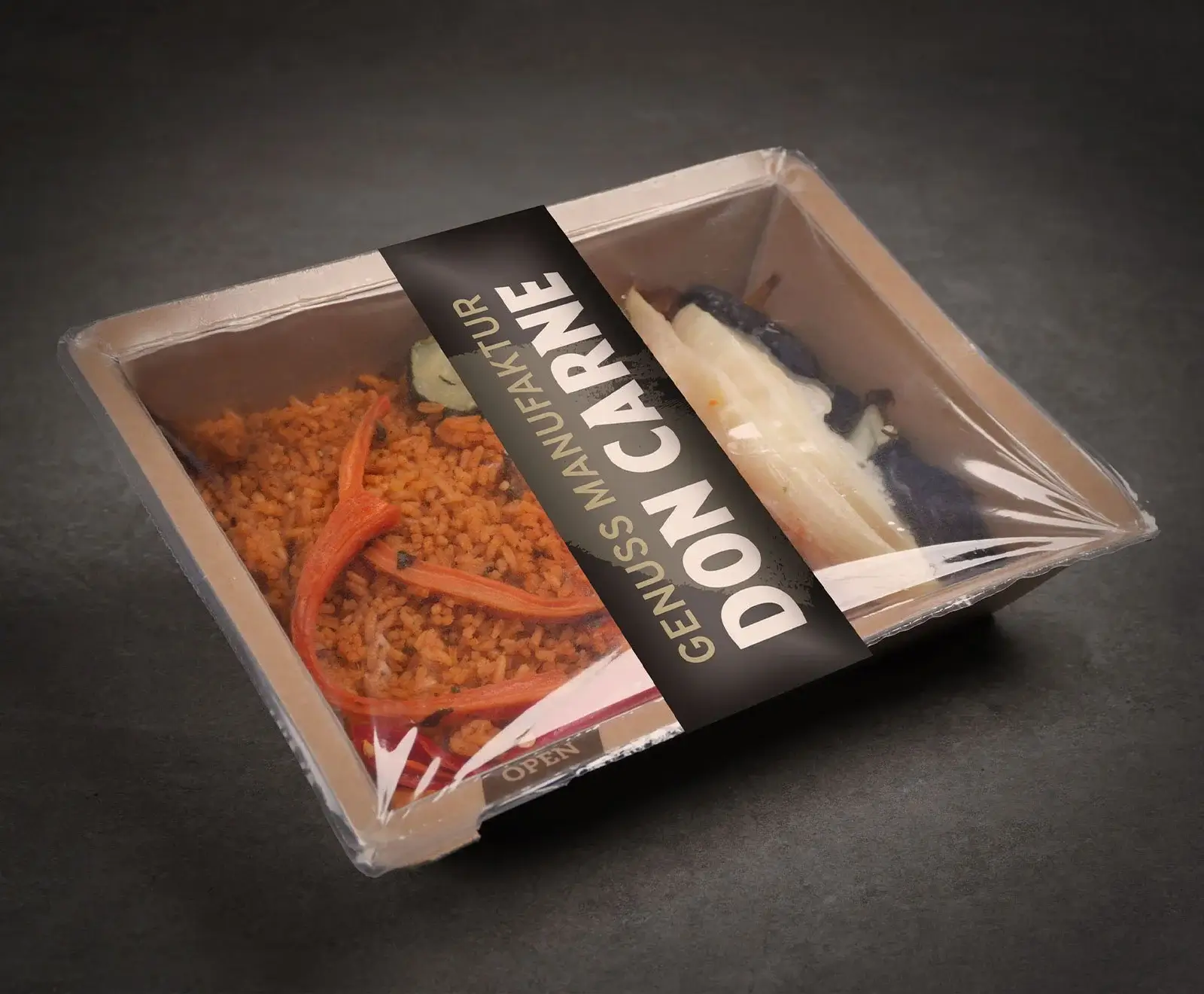 Eine rechteckige Fertigmahlzeit in einer braunen Schale mit Klarsichtdeckel, auf dunklem Untergrund. Sie enthält orangefarbenen Reis mit Karottenstreifen und eine Beilage aus weißen und dunklen Stücken. Ein schwarzes Etikett mit „GENUSS MANUFAKTUR DO