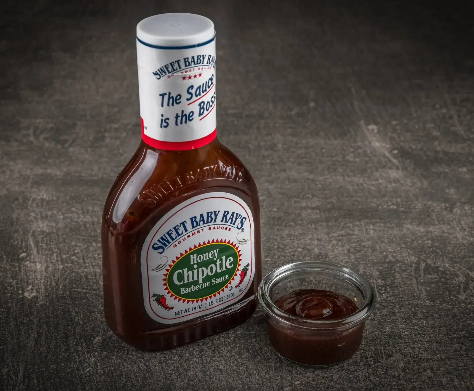 Eine Flasche Sweet Baby Ray's Honey Chipotle Barbecue Sauce steht auf dunklem Untergrund, daneben ein kleines Glas mit der dunklen, glänzenden Sauce. Das Etikett ist deutlich lesbar.