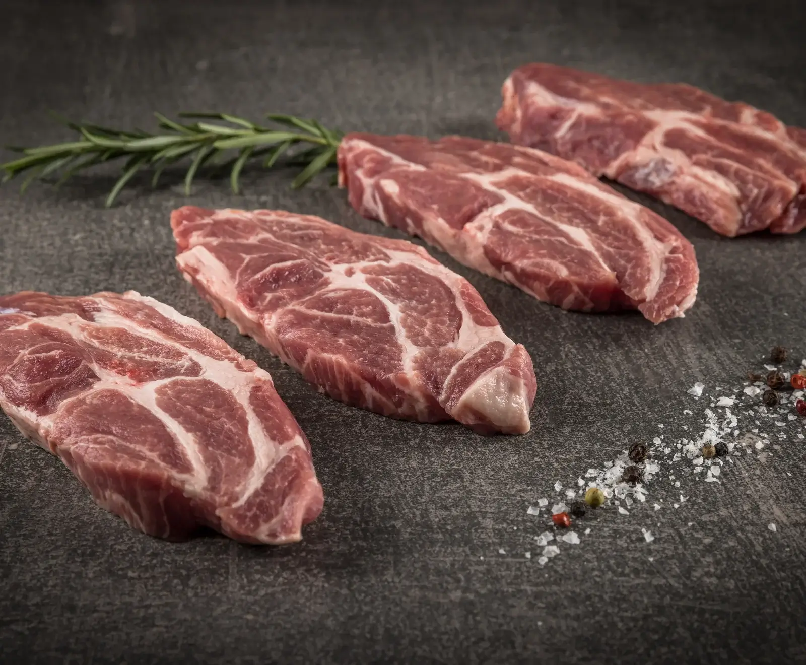 Rohe Schweinenackensteaks mit weißer Marmorierung, dazu Rosmarin, Salz und bunte Pfefferkörner auf dunkler Fläche.