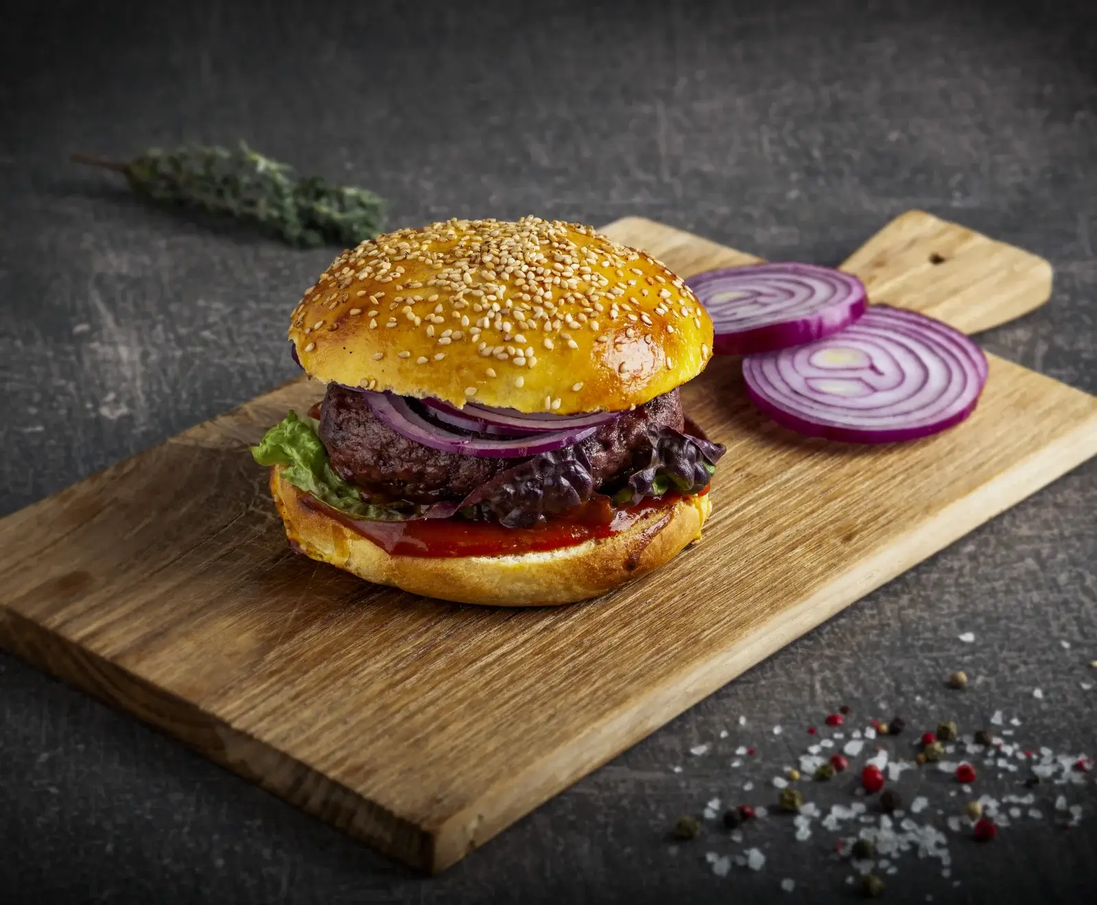 Ein appetitlicher Burger mit Sesambrötchen, dickem Patty, roten Zwiebelringen, Salat und Sauce auf einem Holzbrett. Daneben liegen Zwiebelscheiben, Kräuter und Gewürze auf dunklem Hintergrund.