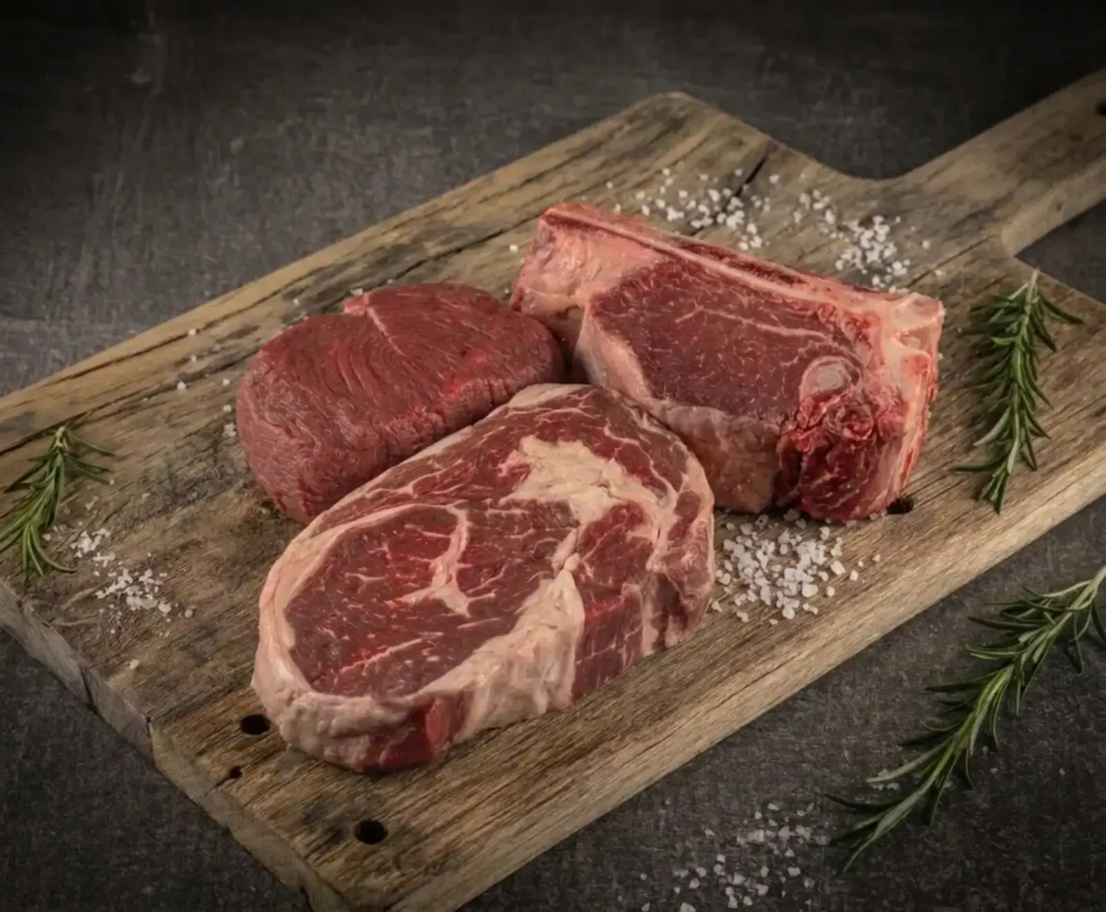Drei rohe Fleischstücke – ein Filet, ein marmoriertes Ribeye und ein Knochensteak – liegen auf einem rustikalen Holzbrett, umgeben von grobem Meersalz und frischen Rosmarinzweigen auf dunklem Hintergrund.