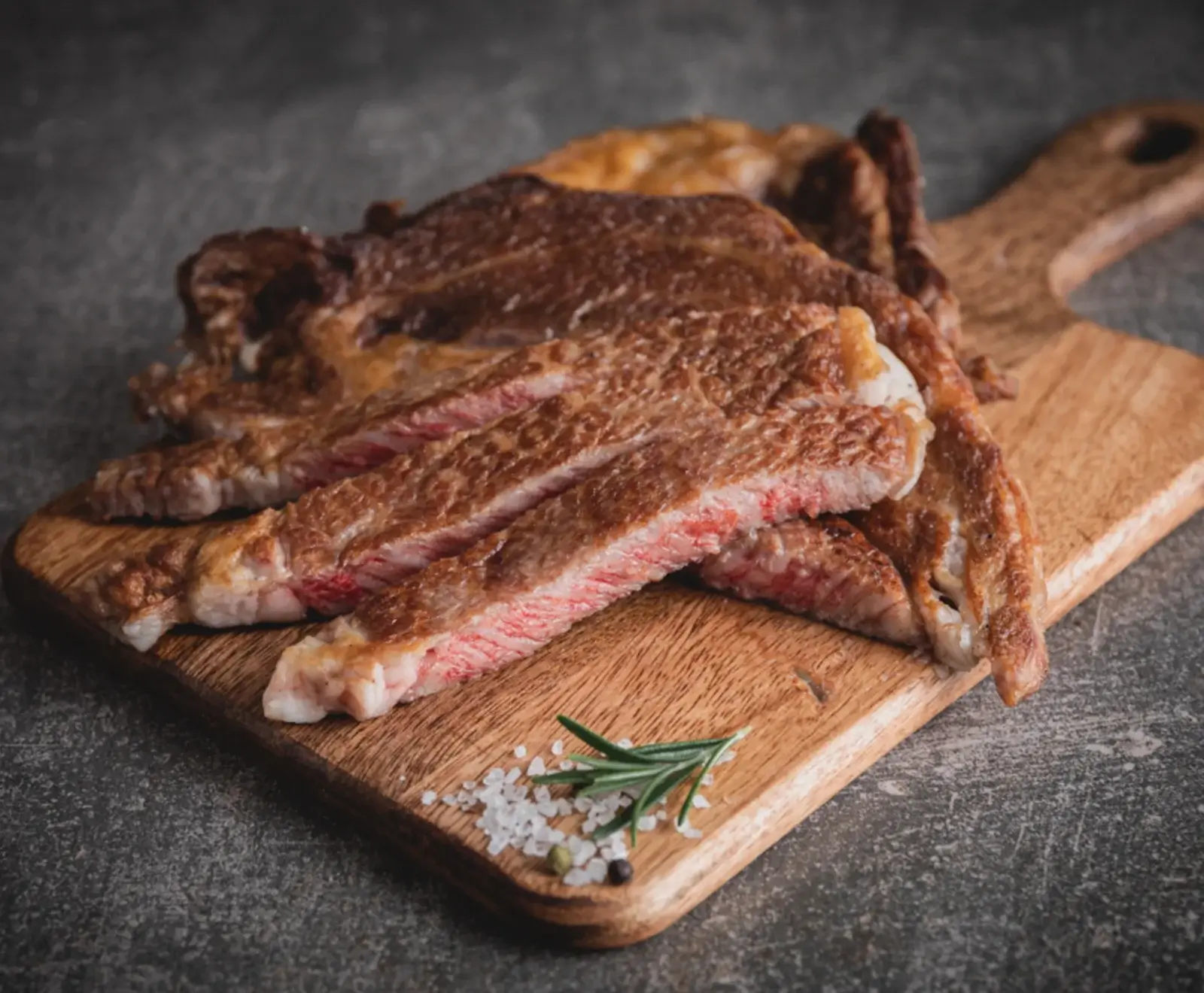 Nahaufnahme von geschnittenem, medium-rare gegartem Steak auf einem Holzschneidebrett, garniert mit grobem Salz und einem Rosmarinzweig auf dunklem Hintergrund.