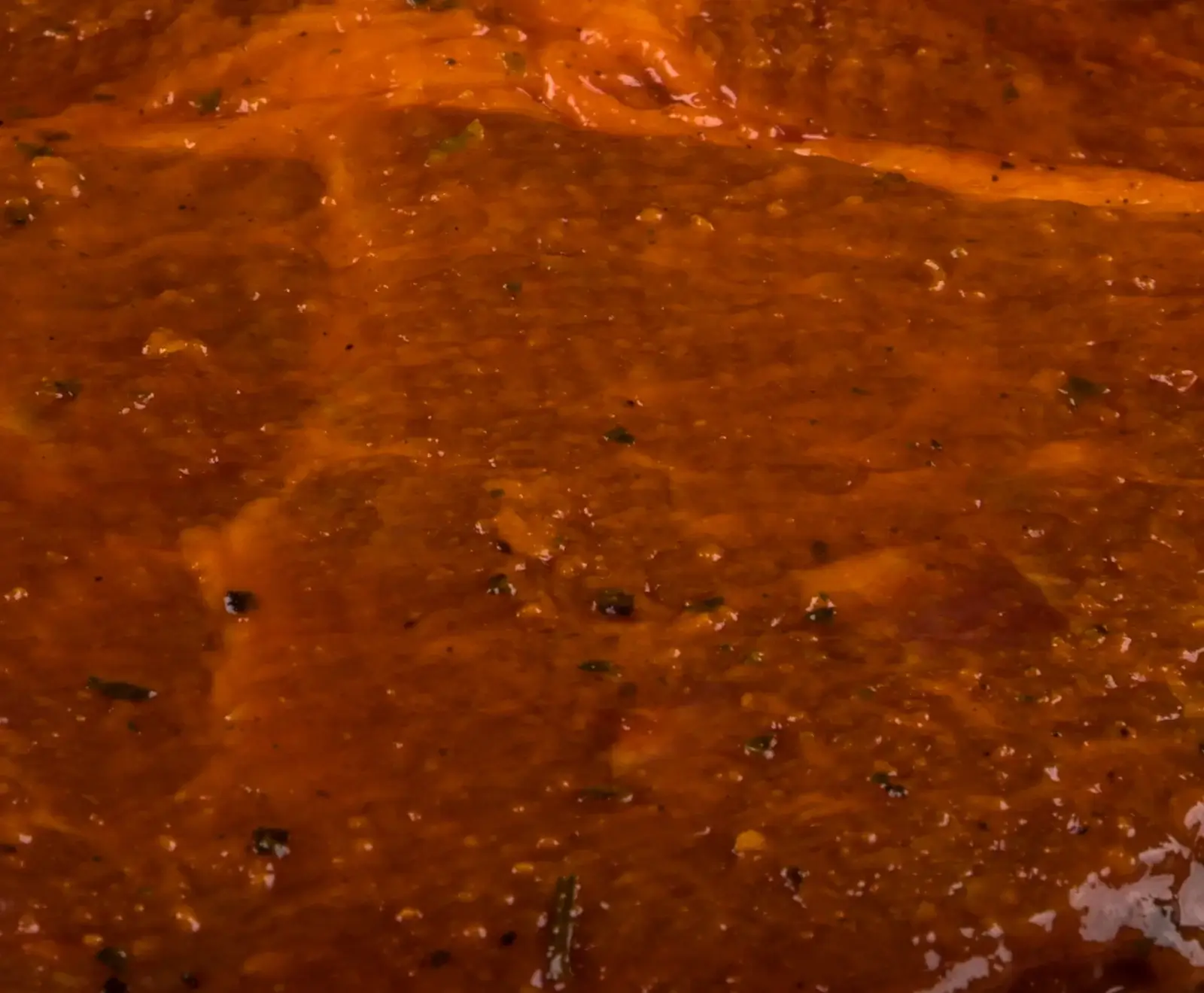 Nahaufnahme von rohem Fleisch, vollständig bedeckt mit einer glänzenden, rötlich-orangen Marinade, in der kleine grüne Kräuter und dunkle Gewürzpartikel sichtbar sind.