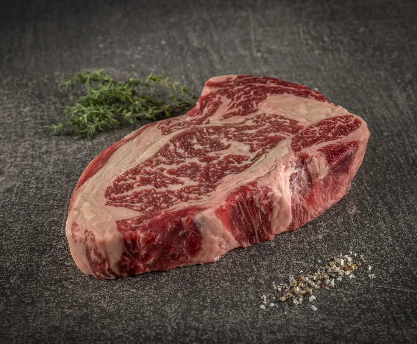 Ein rohes, stark marmoriertes Rindersteak liegt auf dunklem Untergrund, daneben frischer Thymian und eine Prise grobes Salz und Pfeffer.