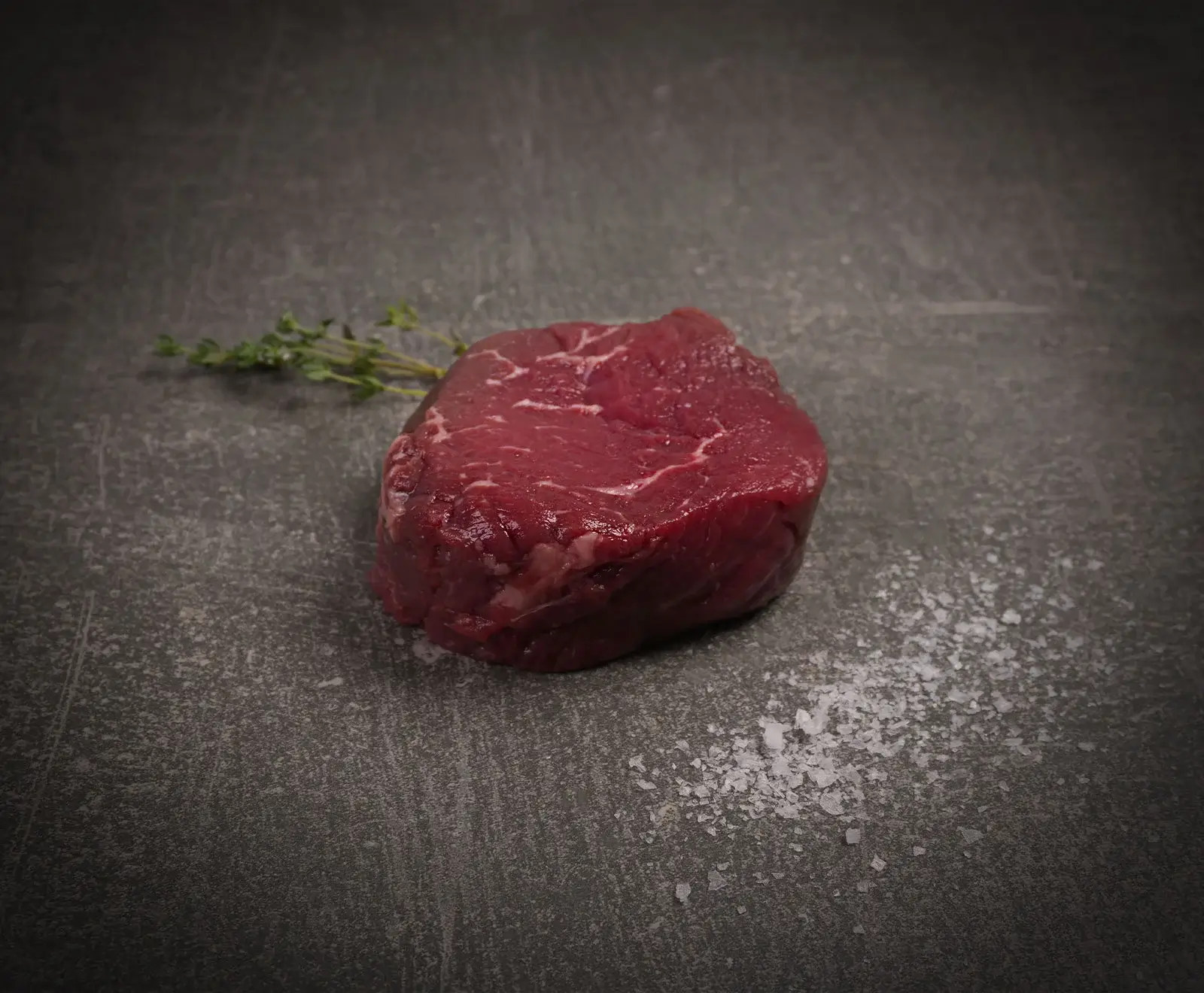 Ein rohes, rotes Rinderfiletsteak liegt auf einer dunklen, strukturierten Oberfläche, daneben ein frischer Thymianzweig und grobe Salzkristalle.