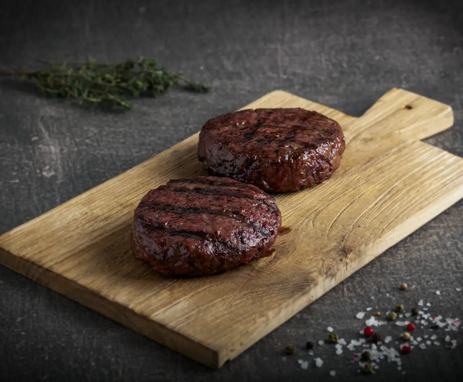 Zwei saftige, gegrillte Burger-Patties mit deutlichen Grillstreifen liegen auf einem rustikalen Holzschneidebrett. Im Hintergrund ist ein Zweig frischer Thymian, im Vordergrund grobes Salz und bunte Pfefferkörner auf dunklem Untergrund.