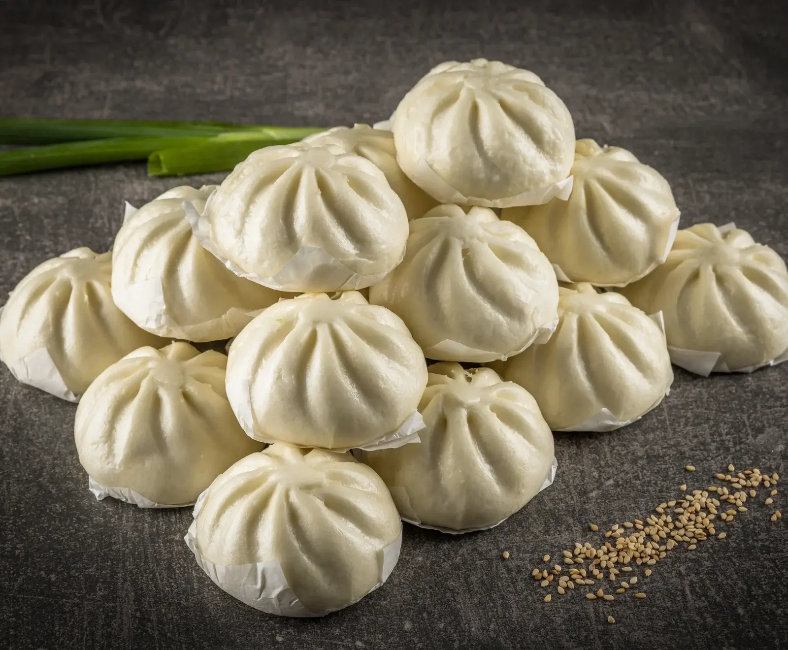 Ein Haufen weißer, gedämpfter Baozi-Brötchen auf dunklem Hintergrund, mit Frühlingszwiebeln und Sesamsamen.
