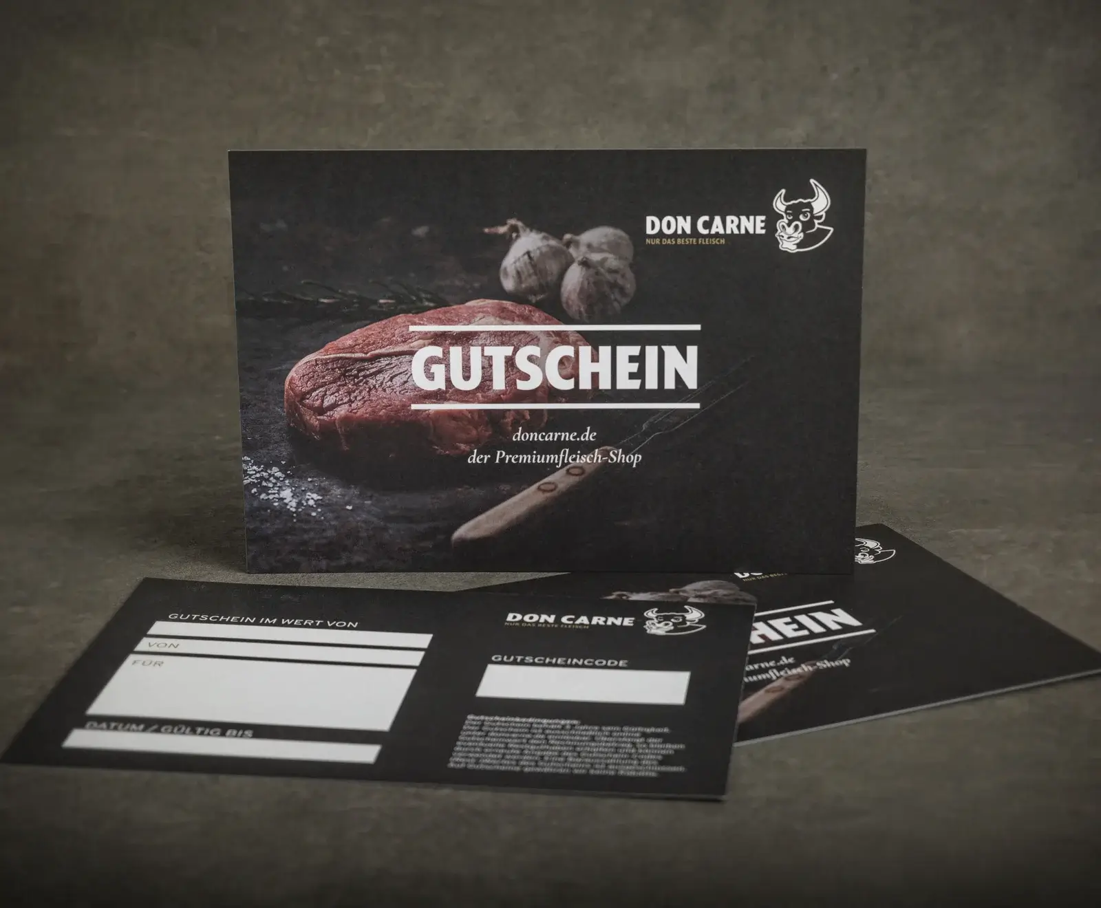 Ein Don Carne Gutschein mit dunklem Design, der ein rohes Steak, Knoblauch und ein Messer zeigt. Im Vordergrund liegen weitere Gutscheinkarten mit Feldern für Wert, Empfänger und Gutscheincode.