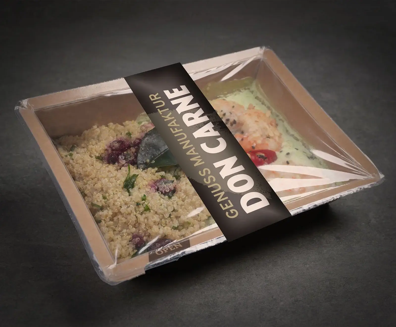 Ein rechteckiger, transparent versiegelter Essensbehälter mit Quinoa, Kräutern und Beeren auf der linken Seite und Garnelen mit grüner Soße und Tomate auf der rechten Seite. Ein schwarzes Etikett mit "DON CARNE GENUSS MANUFAKTUR" ist diagonal angebrac