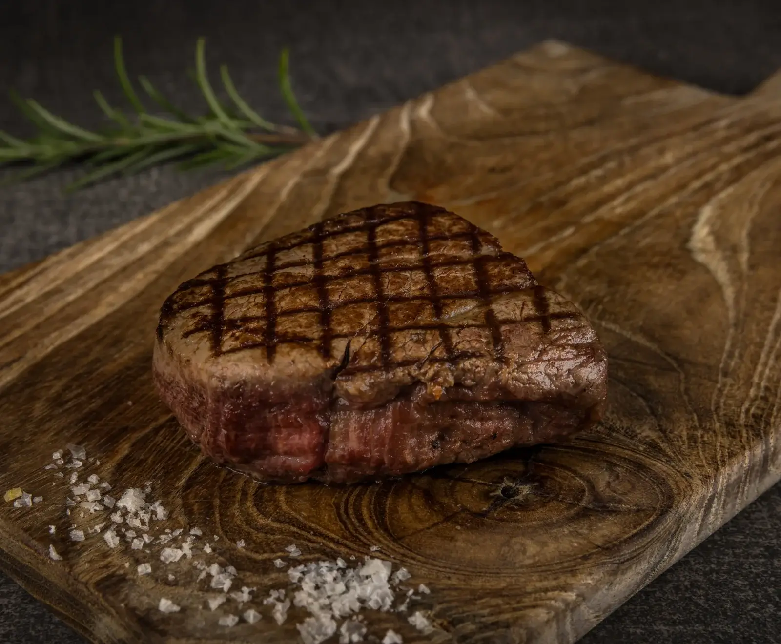 Ein perfekt gegrilltes, dickes Rinderfiletsteak mit deutlichen Grillstreifen liegt auf einem rustikalen Holzbrett. Grobes Meersalz und ein Rosmarinzweig sind daneben platziert. Das Steak ist medium-rare.