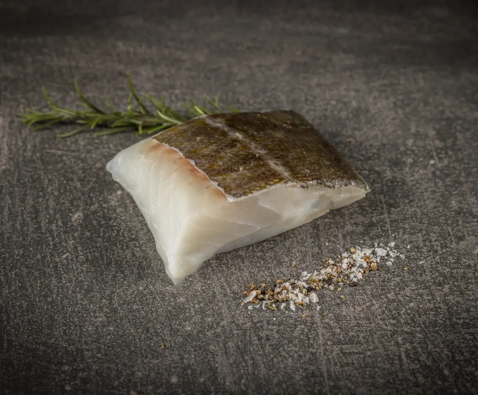 Ein rohes Fischfilet mit Haut, daneben ein Rosmarinzweig und eine Prise grobes Salz und Pfeffer, auf dunklem Steinhintergrund.