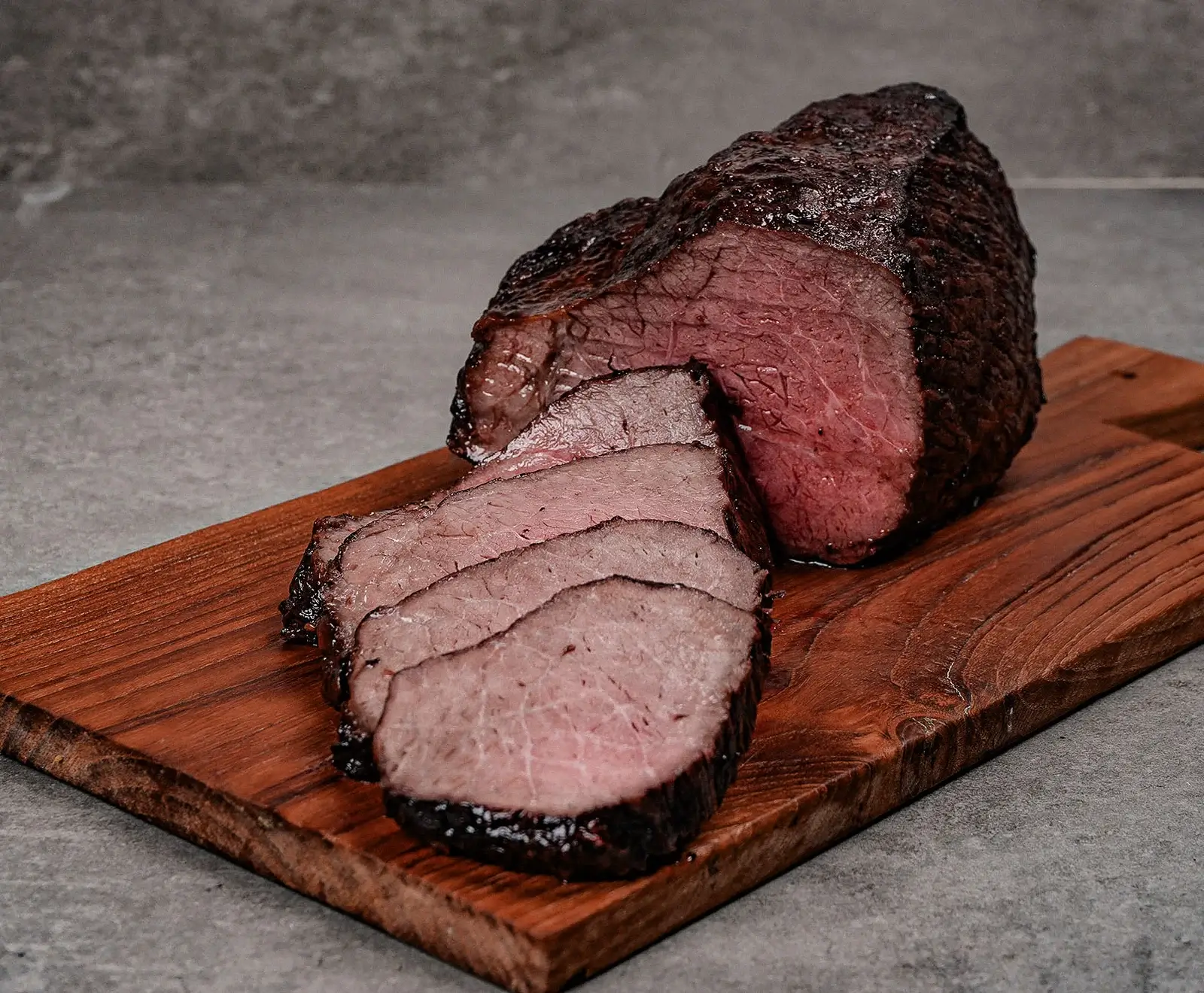 Ein angeschnittener, rosa gebratener Rinderbraten mit dunkler Kruste liegt auf einem rustikalen Holzschneidebrett. Mehrere Scheiben sind daneben platziert, der Hintergrund ist grau.