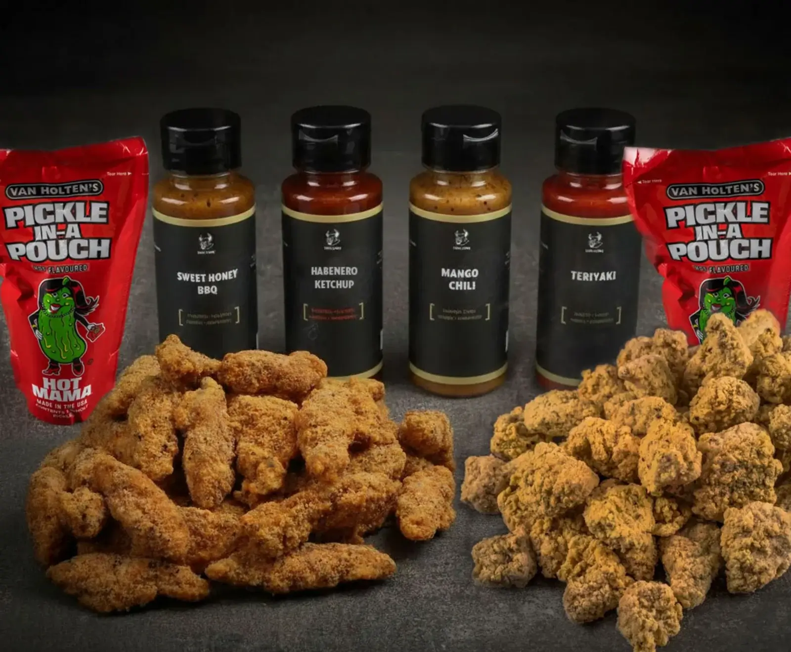 Zwei Haufen frittierter Hähnchenteile, umgeben von vier Saucenflaschen (Sweet Honey BBQ, Habanero Ketchup, Mango Chili, Teriyaki) und zwei roten Van Holten's Pickle In A Pouch 'Hot Mama' Beuteln auf dunklem Hintergrund.