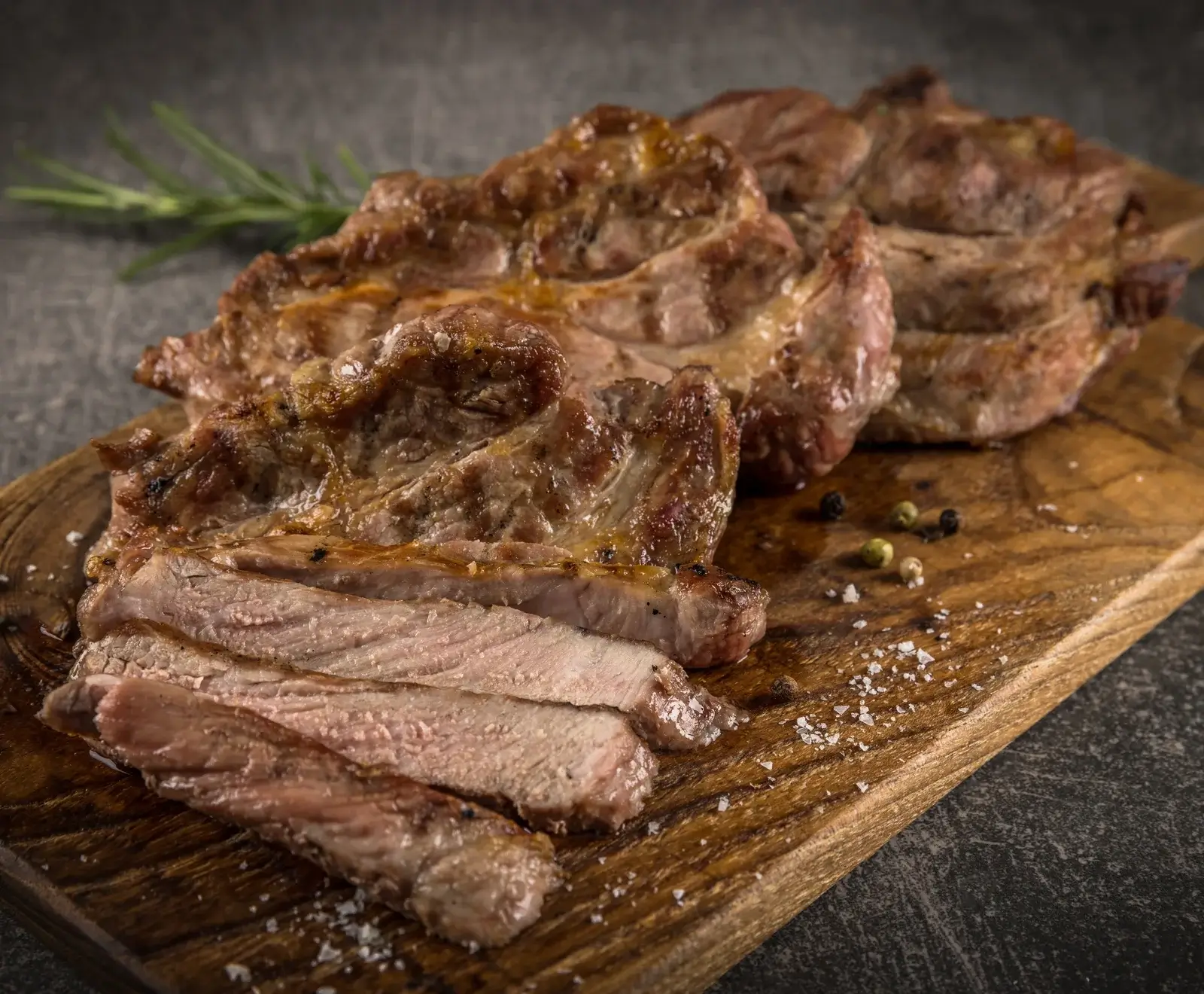 Nahaufnahme von drei goldbraun gegrillten Schweinenackensteaks auf einem Holzschneidebrett. Das vorderste Steak ist in dicke Scheiben geschnitten und zeigt zartrosa Fleisch. Grobes Salz, Pfefferkörner und Rosmarin sind sichtbar.