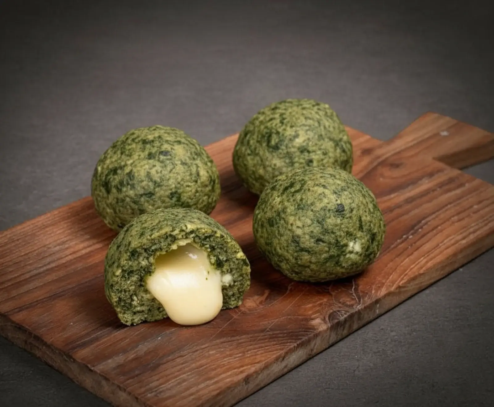 Vier grüne, runde Spinat-Käse-Bällchen auf einem Holzbrett. Eines ist halbiert und zeigt eine cremige, geschmolzene Käsefüllung.