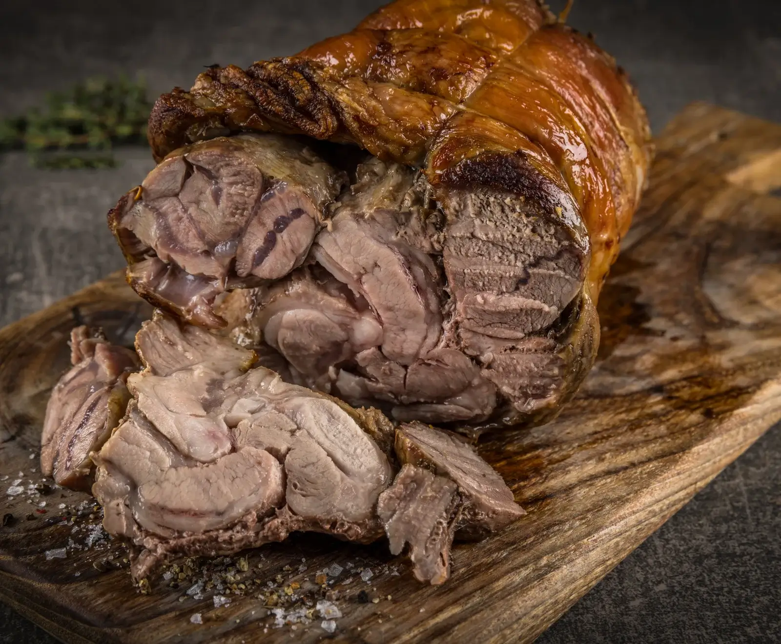 Ein großes Stück knusprig gebratener Schweinebraten mit goldbrauner Kruste, teilweise aufgeschnitten, um das zarte, saftige Fleisch zu zeigen. Es liegt auf einem rustikalen Holzbrett mit grobem Salz und Pfeffer.