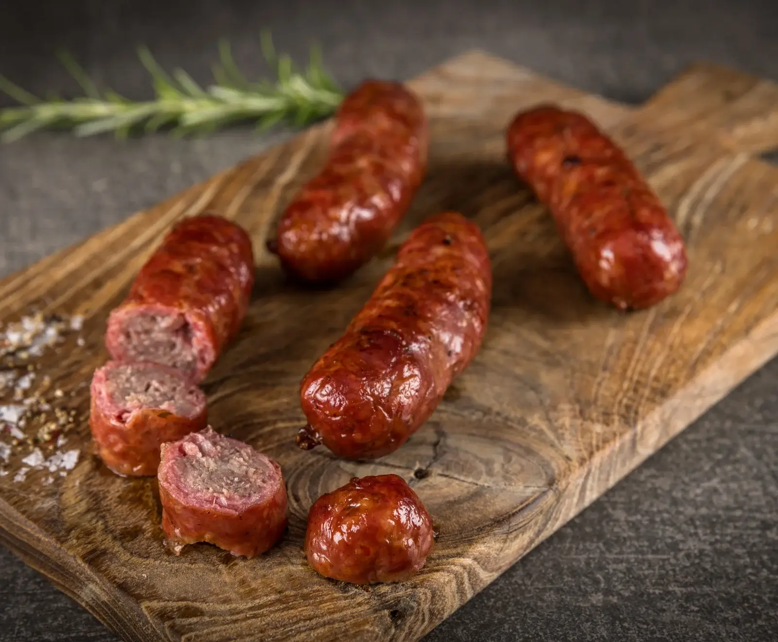 Mehrere gebratene Würste auf einem Holzbrett, eine davon in Scheiben geschnitten, mit grobem Salz und Rosmarin im Hintergrund. Die Würste haben eine glänzende, rötlich-braune Farbe.