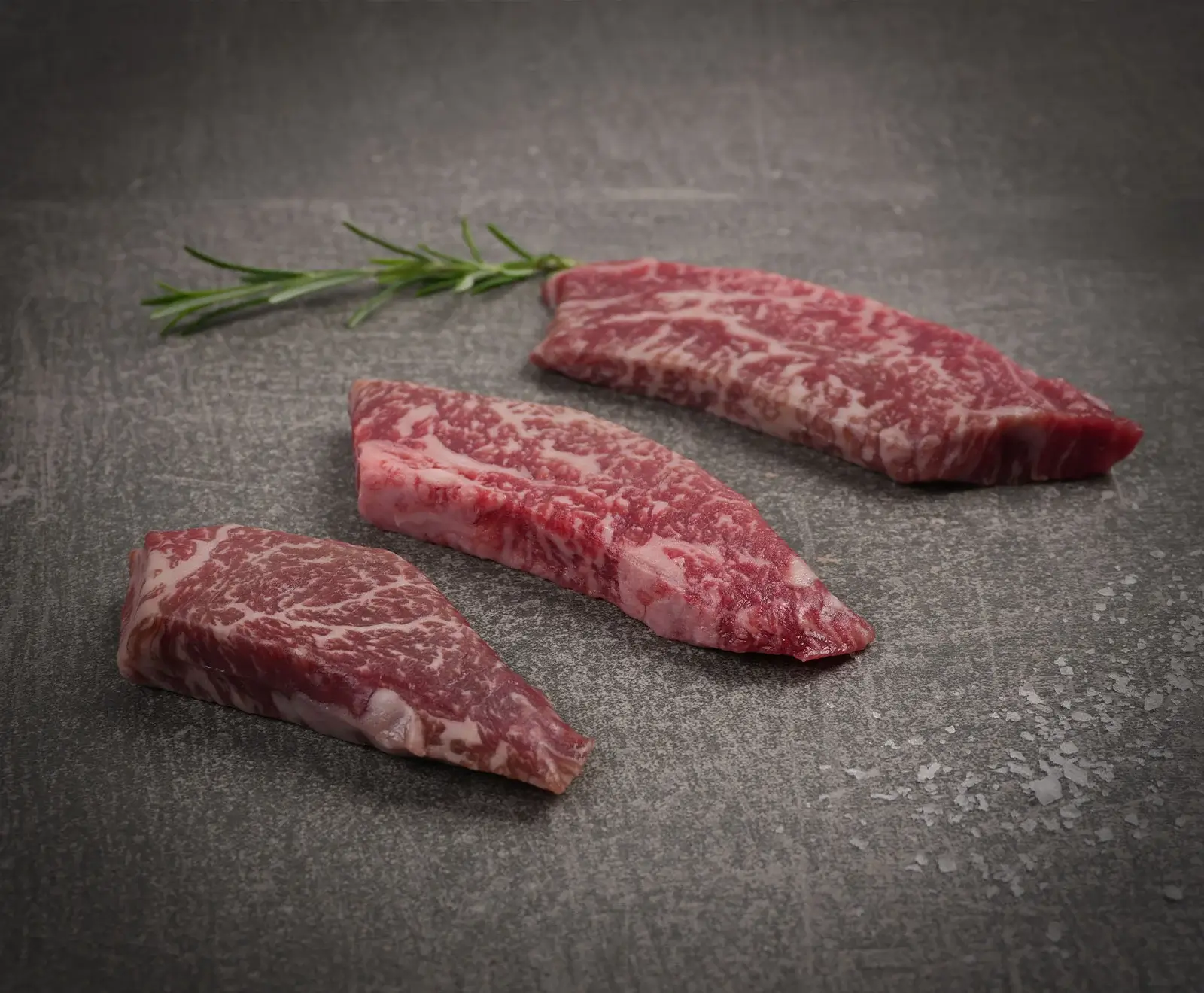 Drei rohe, stark marmorierte Rindfleischstücke liegen diagonal auf einer dunklen, strukturierten Oberfläche. Ein Rosmarinzweig und grobe Salzkristalle sind daneben platziert.